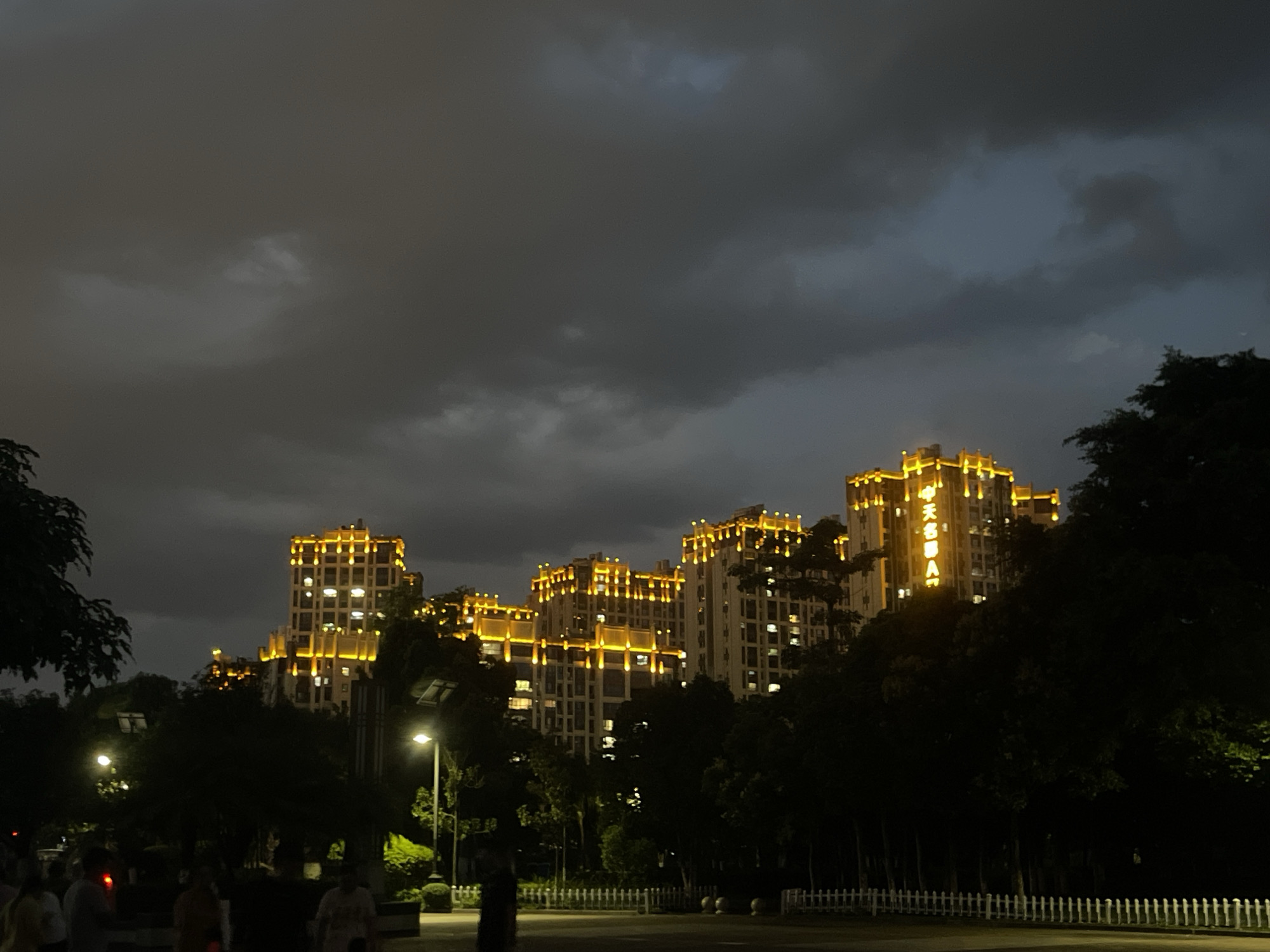 The image depicts a nighttime cityscape with several multi-story buildings illuminated by bright yellow lights. The buildings are modern in design, featuring numerous windows and architectural details that are highlighted by the lighting. Trees and foliage are visible in the foreground and between the buildings, casting shadows and creating a sense of depth. The sky is dark and cloudy, suggesting a twilight or night scene. Streetlights are also present, casting pools of light on the ground. A white picket fence runs along the bottom of the frame, bordering a pathway or road. Several indistinct figures of people are visible in the lower left and center portions of the image, appearing to be walking or standing. The overall atmosphere is one of an urban environment at night, with the illuminated buildings serving as a focal point against the dark sky.