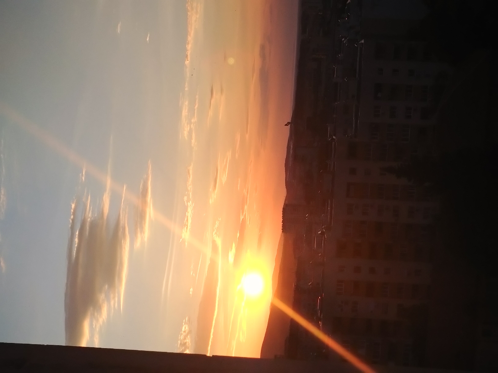 The image captures a vibrant sunset over a section of Stara Zagora, Bulgaria, viewed from an elevated position. The sky is a blend of blues and oranges, with the sun positioned near the horizon, casting bright rays. Clouds are scattered across the sky, reflecting the colors of the sunset. In the right portion of the image, the silhouette of a multi-story building is visible against the bright sky. The building appears to be a residential block, as indicated by the rows of windows. The foreground is dark, showing the edge of a structure where the photo was taken.