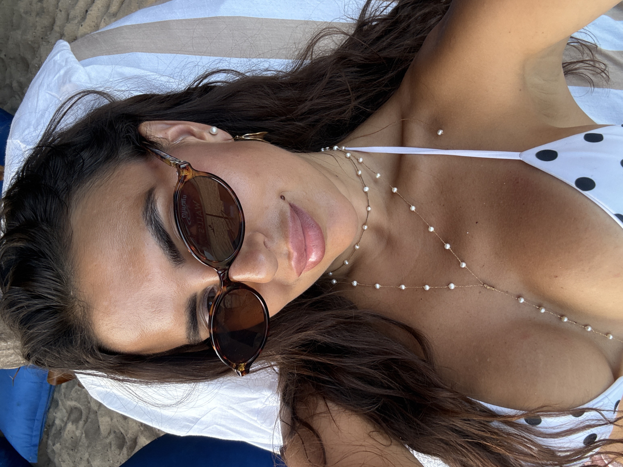 A woman lies supine on a white towel, presumably on a beach. She is wearing sunglasses with tortoise shell frames and a white bikini top with black polka dots. A delicate gold necklace with small pearls drapes across her chest. Her hair is long, dark brown, and partially spread across the towel. The image appears to be taken in Kuta Selatan, Indonesia, based on the beach setting and skin tone of the subject.