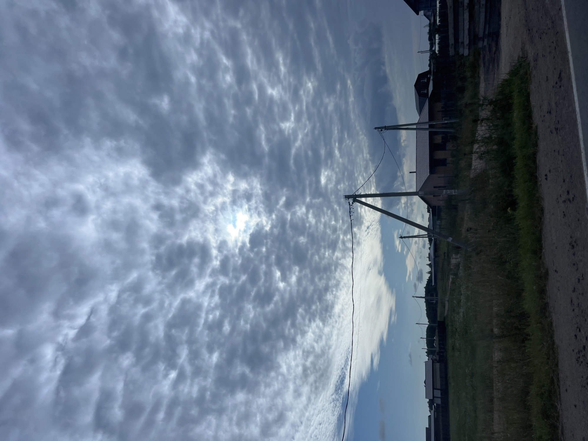 The image captures a view of a rural landscape, likely within the city of Novaya Ropsha, Russia, under a dramatic, cloudy sky. The sky is filled with a textured layer of gray and white clouds, with a patch of brighter sunlight peeking through towards the upper center. In the middle ground, a cluster of buildings is visible, including a house with a red-tiled roof, and other smaller structures and fences. Two wooden utility poles with wires strung between them stand prominently in the foreground, one on the left and another to the right of the house. The ground in the foreground is a rough, dirt road or path, with patches of green grass and weeds growing along its edges. The overall atmosphere is overcast and somewhat muted, with the natural elements of the sky and vegetation dominating the scene.