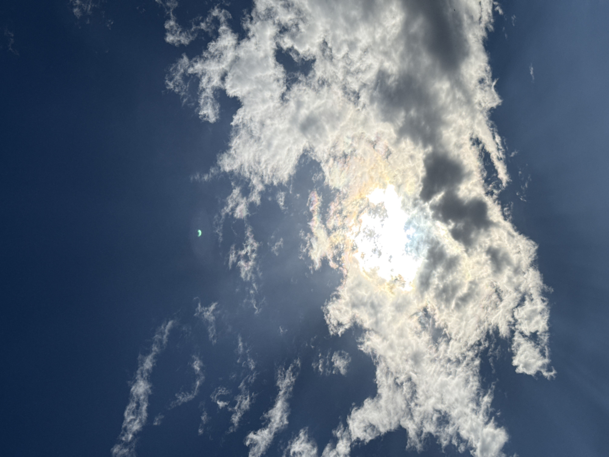 The image shows a daytime sky in Plovdiv, Bulgaria. Predominantly blue, the sky is partially obscured by cumulus clouds. The sun is visible through a break in the clouds, causing bright light and some visible rays. A small green object, possibly a kite or drone, is located in the upper-left quadrant of the image.