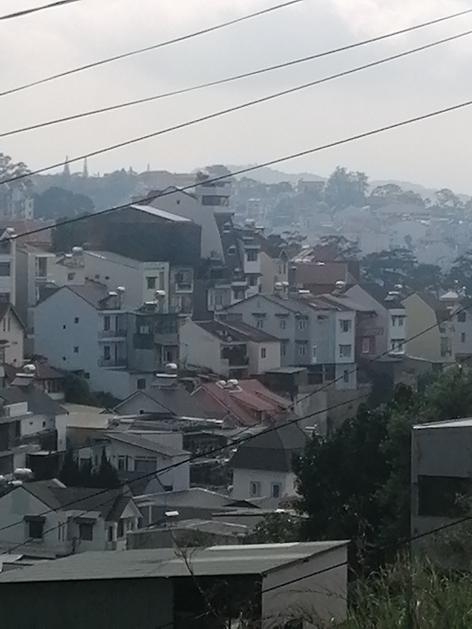The image shows a daytime view of a densely packed residential area in Da Lat, Vietnam, characterized by a variety of buildings of differing heights and architectural styles. Predominantly, the buildings are multi-storied and painted in light colors. Numerous rooftops are visible, some with water heaters or solar panels. In the distance, rolling hills and trees fade into a hazy sky. Power lines are strung across the foreground, partially obscuring the scene. Vegetation is visible in the lower right corner of the image. In the far distance, two spires are visible.