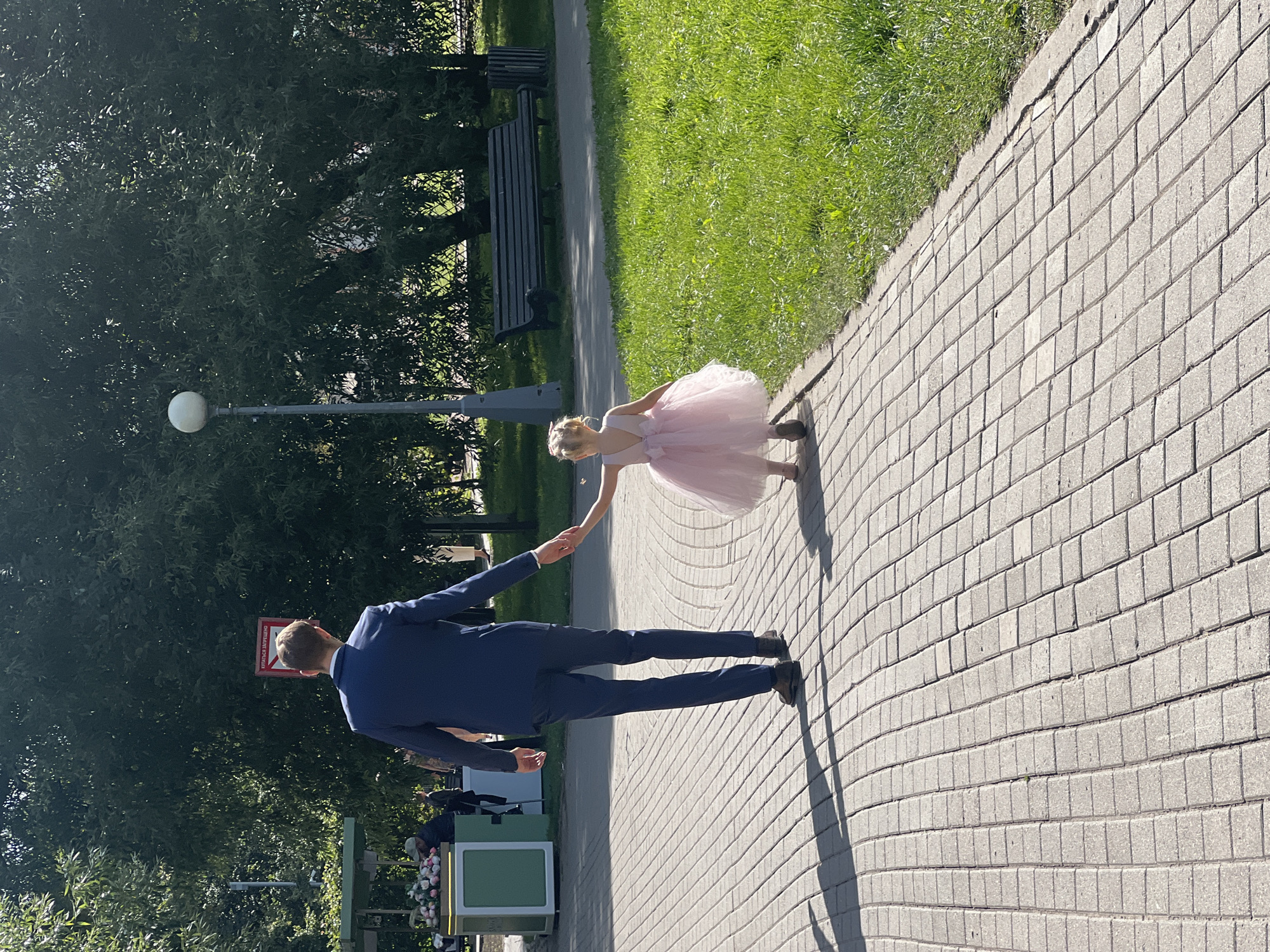 In a city park in Moscow, Russia, a man in a navy blue suit and a young girl in a pink dress are walking on a brick-paved path. The man is holding the girl's hand. The girl is walking away from the camera, and the man is walking next to her, also facing away from the camera. They are in a park with green grass, trees, and a lamppost. In the background, there is a park bench and a trash can. The sun is shining, casting shadows on the path.