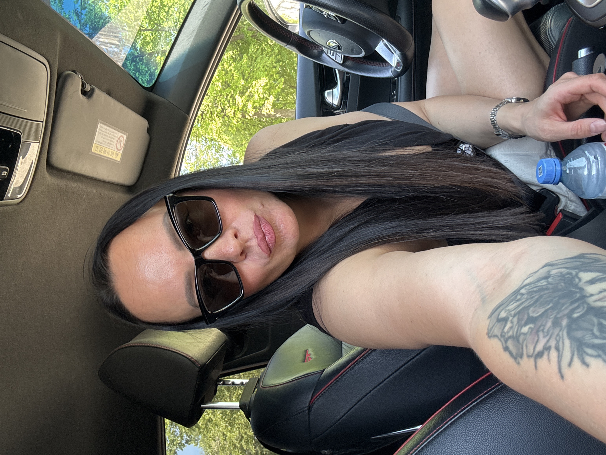 The image is a selfie taken inside a car. A person with long dark hair and sunglasses is visible. The person is wearing a black top and has a tattoo on their right arm. A water bottle is visible next to them. The car's interior includes black leather seats with red stitching and a black steering wheel. The sun visor is visible in the upper left corner. Through the car windows, trees are visible, suggesting an outdoor setting. There is no information that this photo was taken in the city of Chaikovsky, Russia.