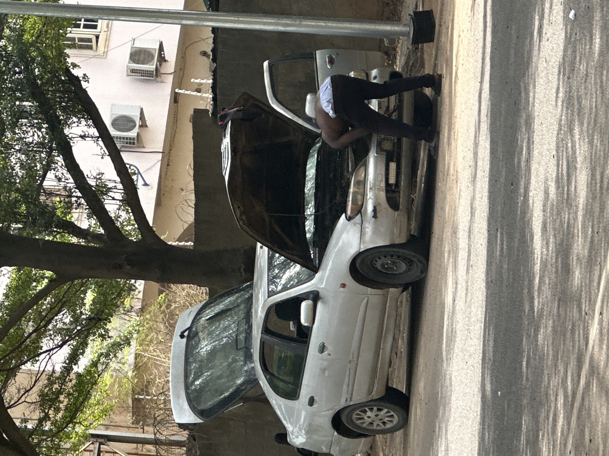 A white car with its hood open is parked on a street in Abuja, Nigeria. A man dressed in dark pants and a white undershirt is bent over the car's engine compartment, appearing to work on the vehicle. The car is positioned near a building with air conditioning units mounted on its exterior. A large tree with dense foliage stands adjacent to the car. Shadows from the tree fall across the road.