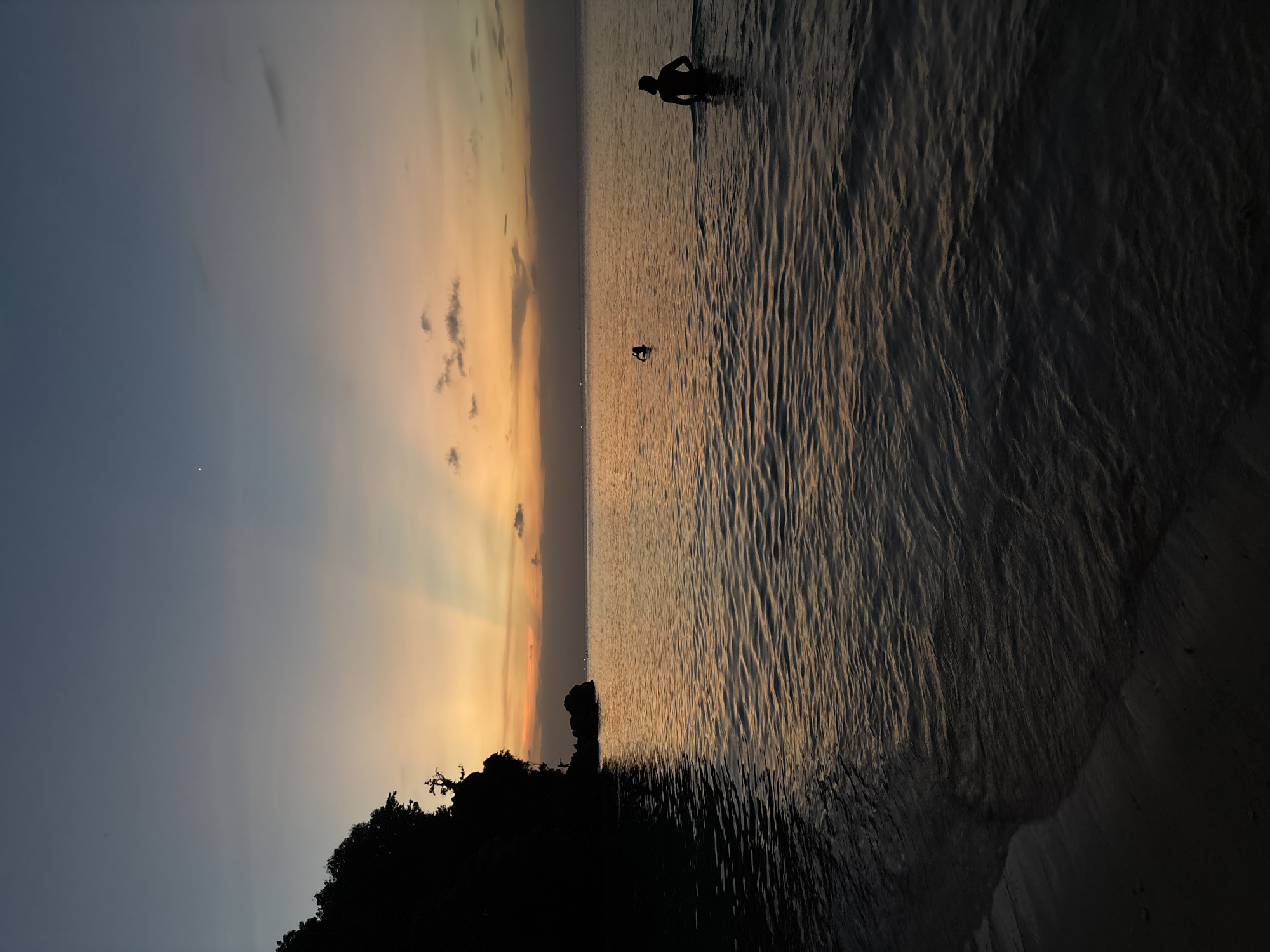 The image depicts a beach scene at sunset, likely in Kuta Selatan, Indonesia. The sky transitions from a deep blue to a golden orange where the sun sets, with visible crepuscular rays. Silhouetted in the water are two people, one appearing to stand, the other farther out, possibly swimming. The sea is rippled, reflecting the sunset colors. In the foreground, dark silhouettes of trees or foliage edge the beach.