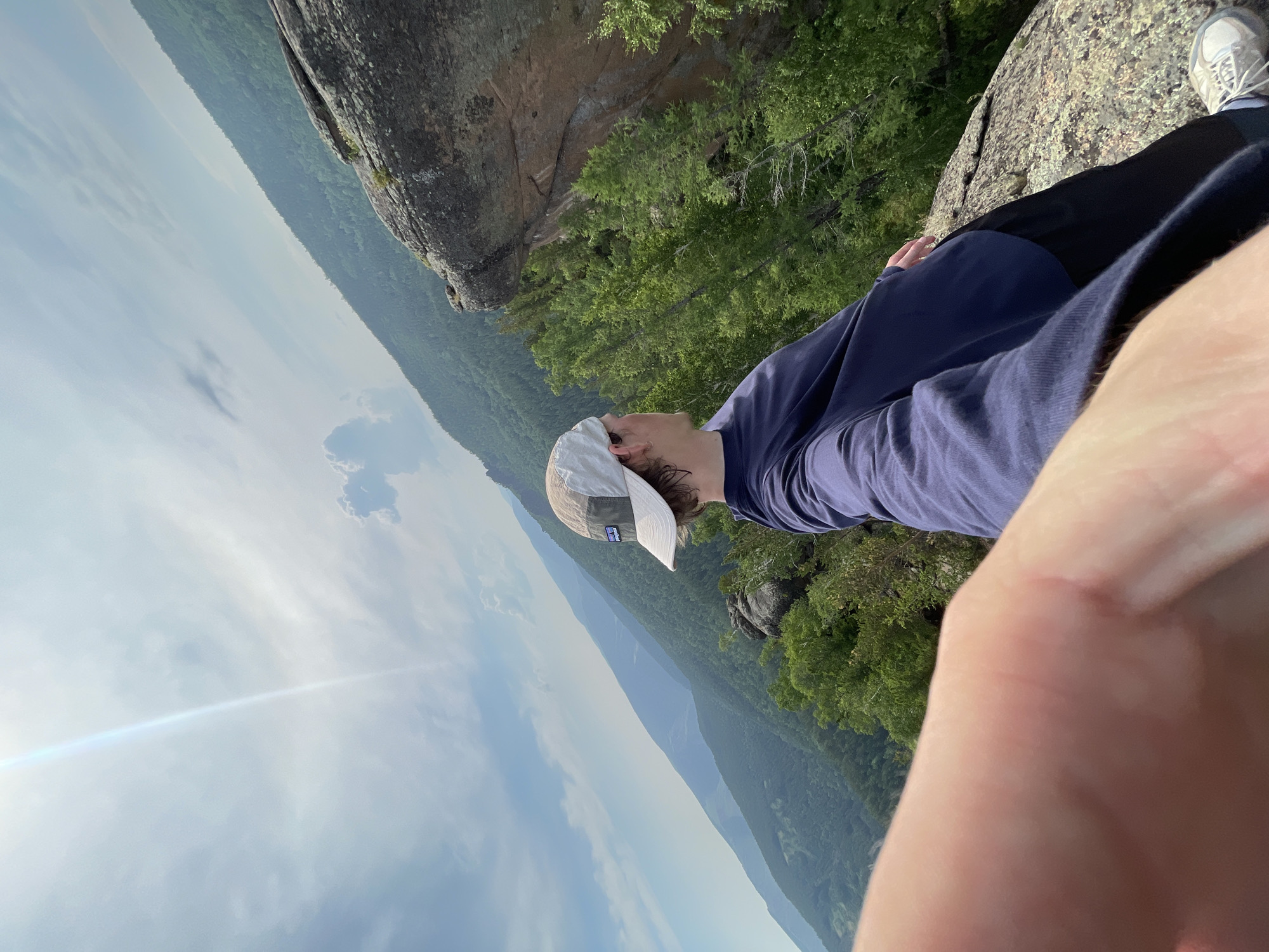 The media file shows a person sitting on a rocky outcrop overlooking a forested valley. The person is wearing a dark blue long-sleeved shirt and black pants, along with a white baseball cap. The setting appears to be a mountainous or hilly area with dense green foliage covering the landscape. The sky is partly cloudy with bright sunlight, suggesting it is daytime. The sun's glare is visible in the frame, indicating a sunny day. The person is looking upwards and to the left, possibly at the scenery or the sky. The perspective of the image is from a low angle, with a significant portion of the foreground occupied by the person's arm and hand.
There is visible text on the person's baseball cap, appearing to be a logo with "PATAGONIA" written on it. The overall mood seems peaceful and contemplative, with the person enjoying the view and the natural surroundings. The terrain suggests a hiking or outdoor recreational activity. Given the location context of Krasnoyarsk, Russia, the scene could be a viewpoint in one of the region's natural parks or mountain ranges. The time of day is likely late morning or early afternoon, based on the sunlight and cloud cover. The weather appears to be pleasant for outdoor activities.