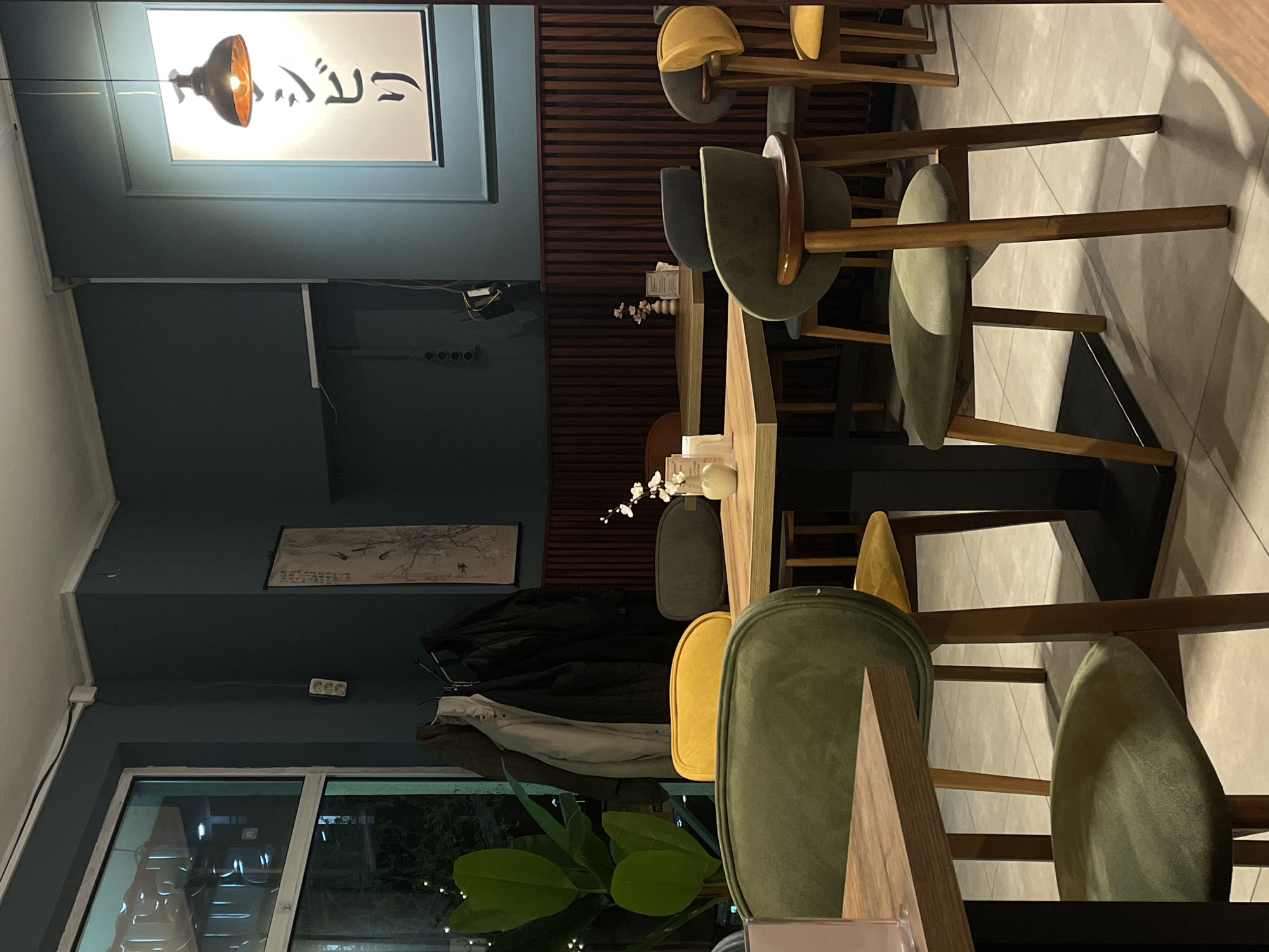 The image depicts the interior of a restaurant or cafe in Batumi, Georgia. There are several chairs with a modern design around a table, and the seats have an olive green and mustard yellow color. A lamp hangs above a framed art piece. In the background, a wall is decorated with dark horizontal slats. A picture with a nature scene is above a row of hooks holding coats. A large window lets in light and shows plants and reflections of light.