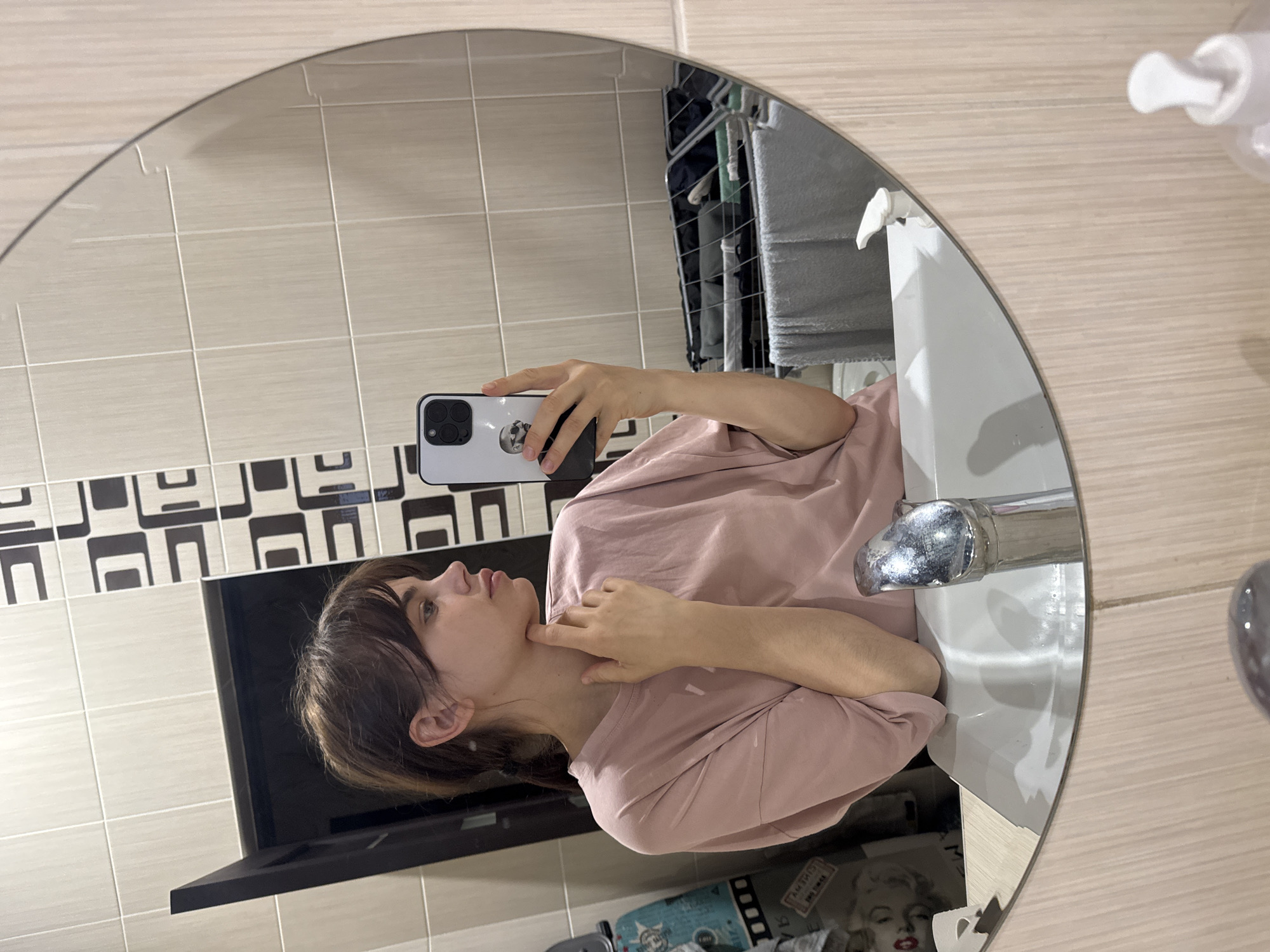A young woman with brown hair and bangs takes a mirror selfie in what appears to be a bathroom. She wears a light pink t-shirt, holding a white smartphone with three visible camera lenses in her right hand. Her left index finger rests on her neck or chin as she looks slightly upwards.
The circular mirror reflects a white sink with a shiny chrome faucet. The bathroom walls are tiled with light beige square tiles, featuring a distinctive black and white patterned border above. In the background reflection, a clothes drying rack with textiles (possibly towels) is visible. To the right, partially seen bottles, likely soap or shampoo dispensers, are reflected on a surface.
Below the main reflection of the sink, there's a glimpse of a blue object with the partially visible text "PARADIS" and "NO. 1". Adjacent to it, a black and white image of Marilyn Monroe is reflected, with the words "MARILYN MONROE" and "NO. 1" also partly legible. The indoor lighting suggests no specific time of day. The woman's expression is neutral and contemplative. The setting is likely a residence in Rostov-on-Don, Russia, based on the provided location context.