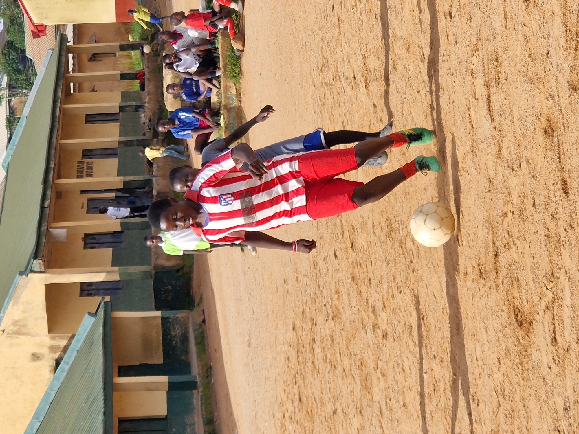 A group of young boys are playing soccer on a dusty field in front of a weathered school building in Kabong, Nigeria. The building, painted yellow and green, appears old and in disrepair. Some boys are actively playing, while others are sitting on a raised step or watching from the sidelines. The ground is made of dry, sandy soil, characteristic of many urban and rural areas in Nigeria. The overall scene depicts a casual game of soccer, a popular pastime among children in Nigeria.