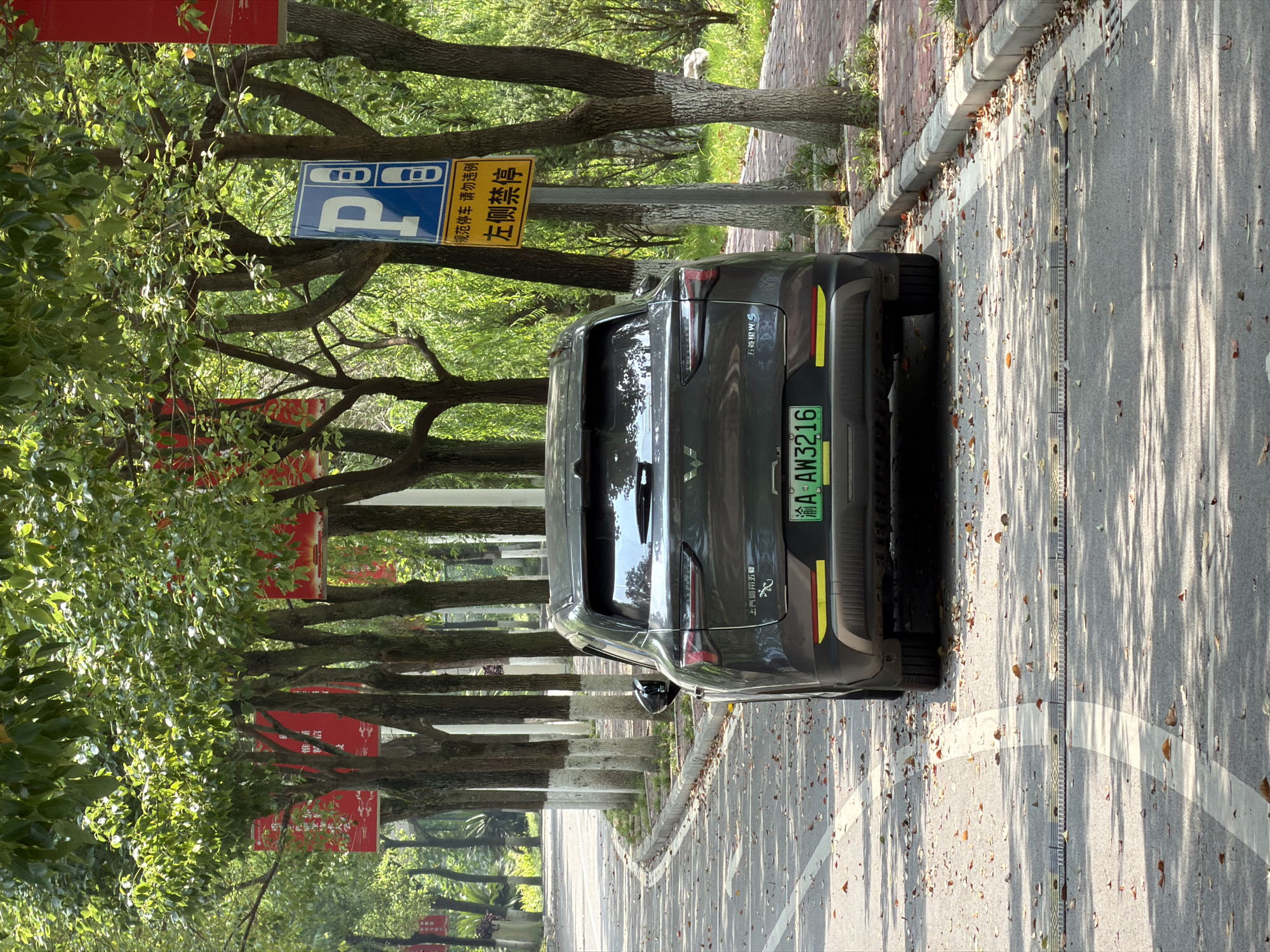 In the city of Jiangjin, China, on a paved road lined with trees and foliage, a dark-colored SUV is parked. The vehicle has a green license plate that reads "AW3216". Adjacent to the road, a blue and white parking sign is visible, indicating "No Parking on the Left Side". The road is partially covered with fallen leaves. The lighting suggests it is daytime, with shadows cast by the trees.