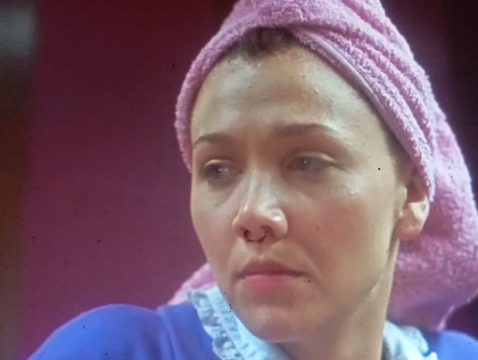 The media file shows a close-up, slightly grainy, medium shot of a woman's face. She is wearing a pink towel wrapped around her head, and a blue garment with white trim around her neck. The background is a blurred, deep pink. The woman's expression is neutral, and she appears to be looking slightly to her left. Her skin is fair, and there are some dark specks visible on her face and in the image itself, possibly dust or imperfections in the recording. Her nose has a distinct septum piercing with a decorative element. The lighting suggests an indoor setting, and the time of day and weather are not discernible. There is no visible text in the media file. The image focuses on the woman's features and attire, with no other objects or people prominently featured. The overall impression is of a candid or posed portrait taken in a domestic setting.
