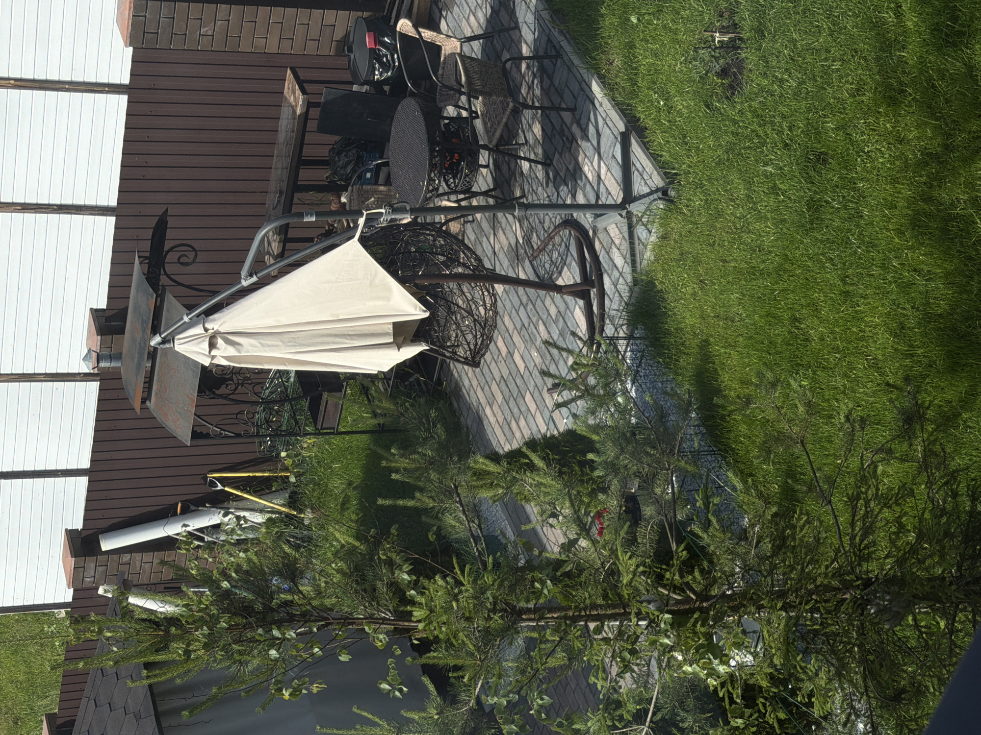 The image depicts an outdoor patio area, likely in a suburban or rural setting. The patio is paved with interlocking bricks and features a round, dark-colored outdoor dining table with chairs. A beige patio umbrella is partially deployed, casting a shadow. To the left, there is a hanging egg chair swing. In the background, a brown wooden fence separates the patio from a white panel fence. A metal barbecue grill is visible near the fence. Several green coniferous trees and lush green grass surround the patio. Various gardening tools, including a shovel and rake, lean against the fence near a downspout. The overall scene suggests a well-maintained outdoor space for relaxation and entertainment.
