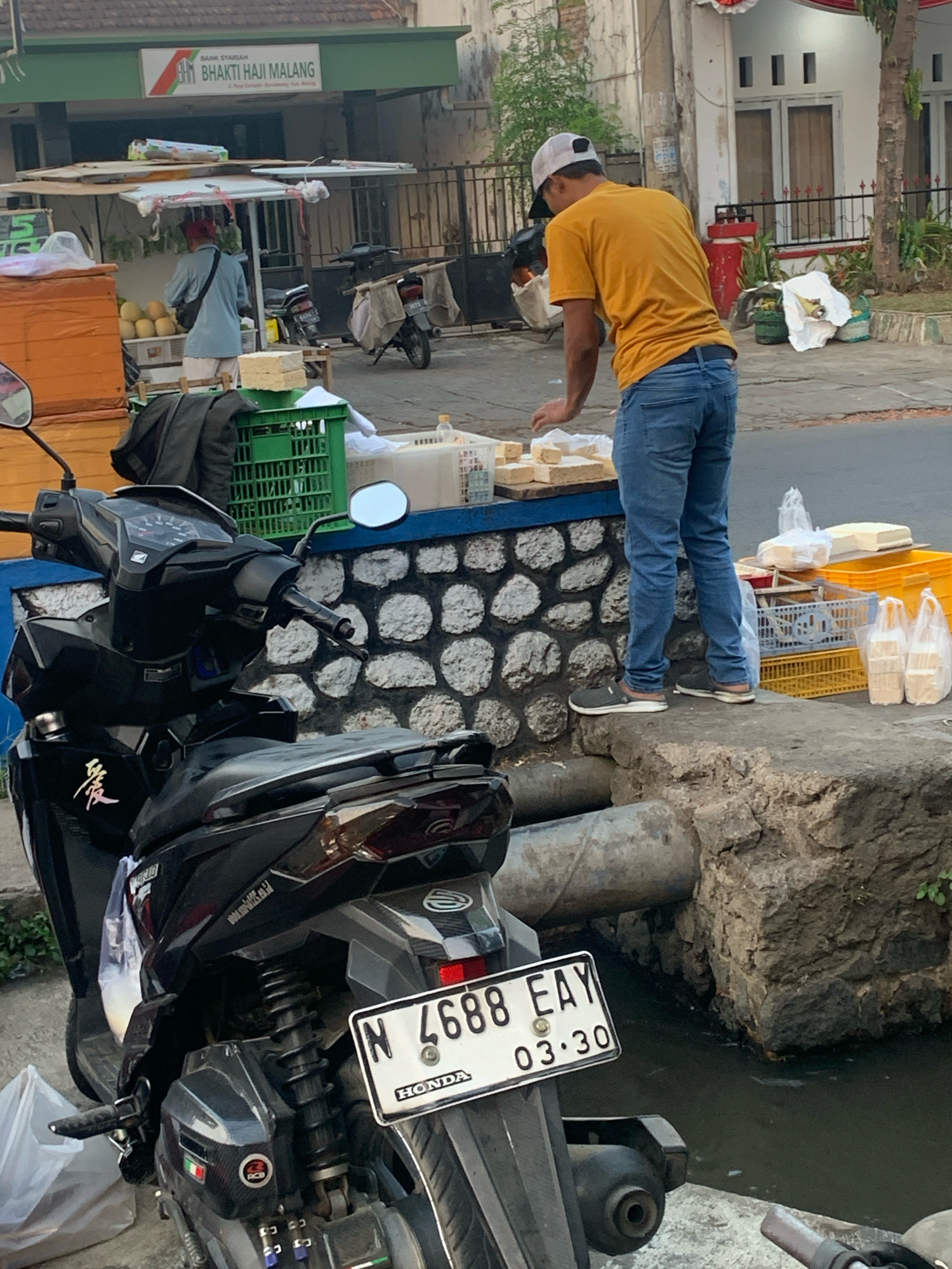 A man wearing a yellow t-shirt, blue jeans, and a white baseball cap is standing on a concrete ledge in front of a stone wall, arranging rectangular food items on a counter. He is facing away from the camera. In the foreground, a black motorcycle is parked. Behind the man, a fruit stall with a red and white canopy is visible, with a person in a blue shirt standing near it. Various crates and plastic bags containing food items are placed on the counter and on the ground. The background shows buildings with signs, including one that reads "BANK SYARIAH BHAKTI HAJI MALANG". The scene appears to be an outdoor market or roadside stall in Bululawang, Indonesia, indicated by the Indonesian script on the bank sign and the presence of a motorcycle with an Indonesian license plate (N 4688 EAY). The overall environment suggests a commercial area with street vendors.