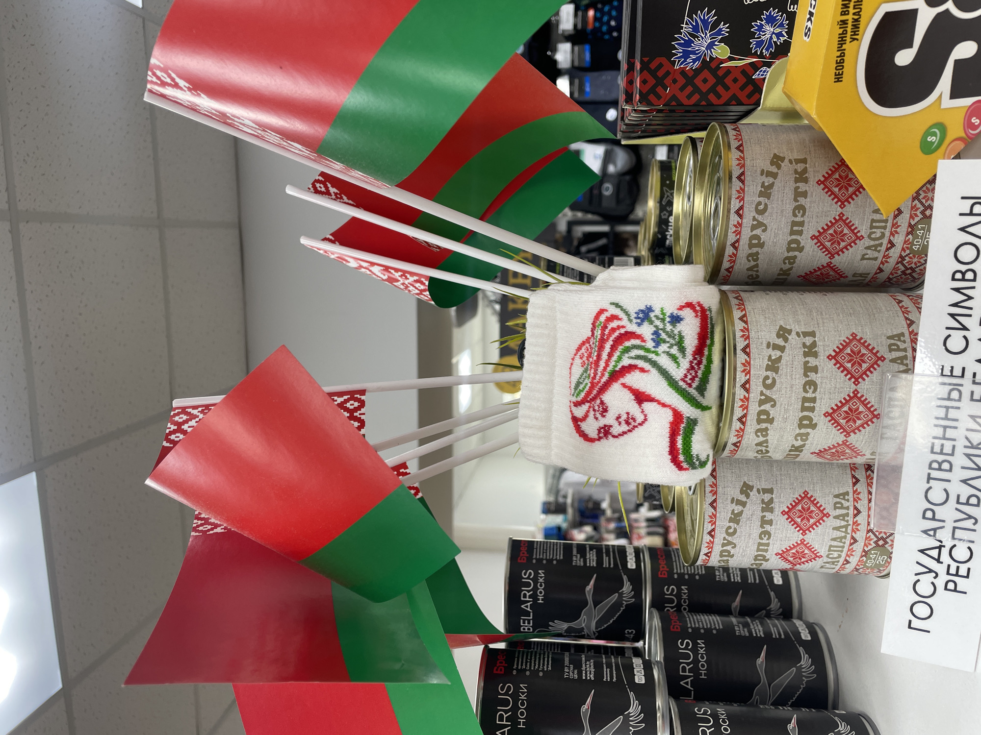 This media file displays a close-up, vertically oriented shot of a retail display, likely in a store in Brest, Belarus. The primary subjects are small Belarusian flags, cans of goods, and packaging for socks. The flags, featuring the red and green colors of Belarus with white ornamental patterns, are clustered together on white poles.
Beneath the flags, stacked cans are visible, wrapped in fabric-like material adorned with traditional Belarusian red and white embroidery patterns. Text on these cans appears to be in Belarusian and indicates "Belarusian carpets" and "Gaspadara" (meaning "hostess" or "mistress of the house" in Belarusian).
Further down, cylindrical packaging for "BELARUS BREST NOSKI" (socks) is stacked. These black cans feature an image of a white stork, a national symbol of Belarus, and text in both Belarusian and English, indicating sock sizes and website information.
To the upper right, a yellow box of Skittles candies is partially visible, along with other stacked retail items. A white label with large black text in Russian reads "GOVERNMENTAL SYMBOLS OF THE REPUBLIC OF BELARUS."
The lighting suggests an indoor setting, typical of a shop or market. The overall impression is a display of national merchandise, highlighting Belarusian culture and products, with a patriotic theme evident through the flags and traditional motifs. The scene is static, with no people or dynamic activity depicted, focusing solely on the products.