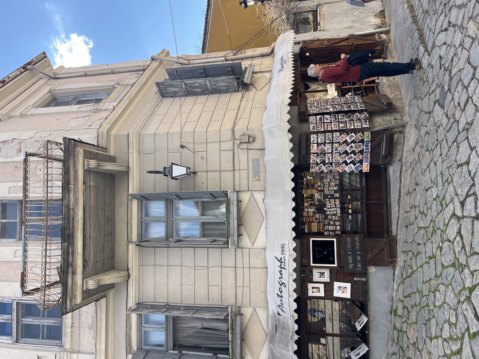The image captures a street scene in Plovdiv, Bulgaria, featuring a building with aging architecture and a photography shop. The building's façade shows signs of wear, with peeling paint, aged windows, and a rusted balcony. The photography shop, identified by a white awning with the word "Autographia since 1994" written on it, has a display of numerous photographs arranged on panels. An elderly man, wearing a red shirt and dark pants, stands in front of the shop, observing the photographs, a black bag hanging from his shoulder. The street is paved with cobblestones, with some grass growing between the stones.
