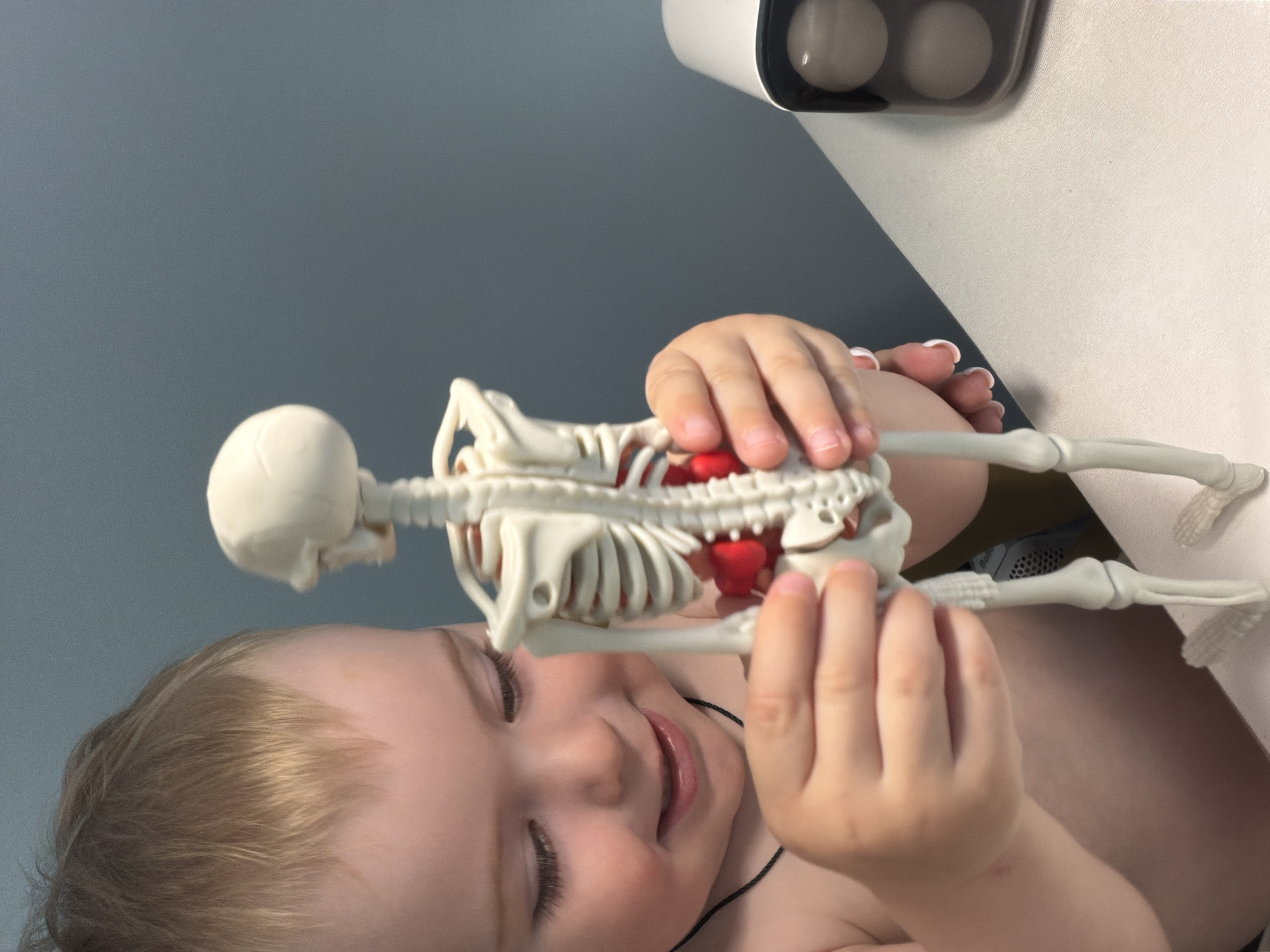 A young child is holding a plastic skeleton toy. The skeleton has a red heart within the rib cage. The child's hands are gripping the skeleton. The background is a grey wall and a white surface with a white object on it. The location is not discernible in the image and cannot be determined to be in Votkinsk, Russia.