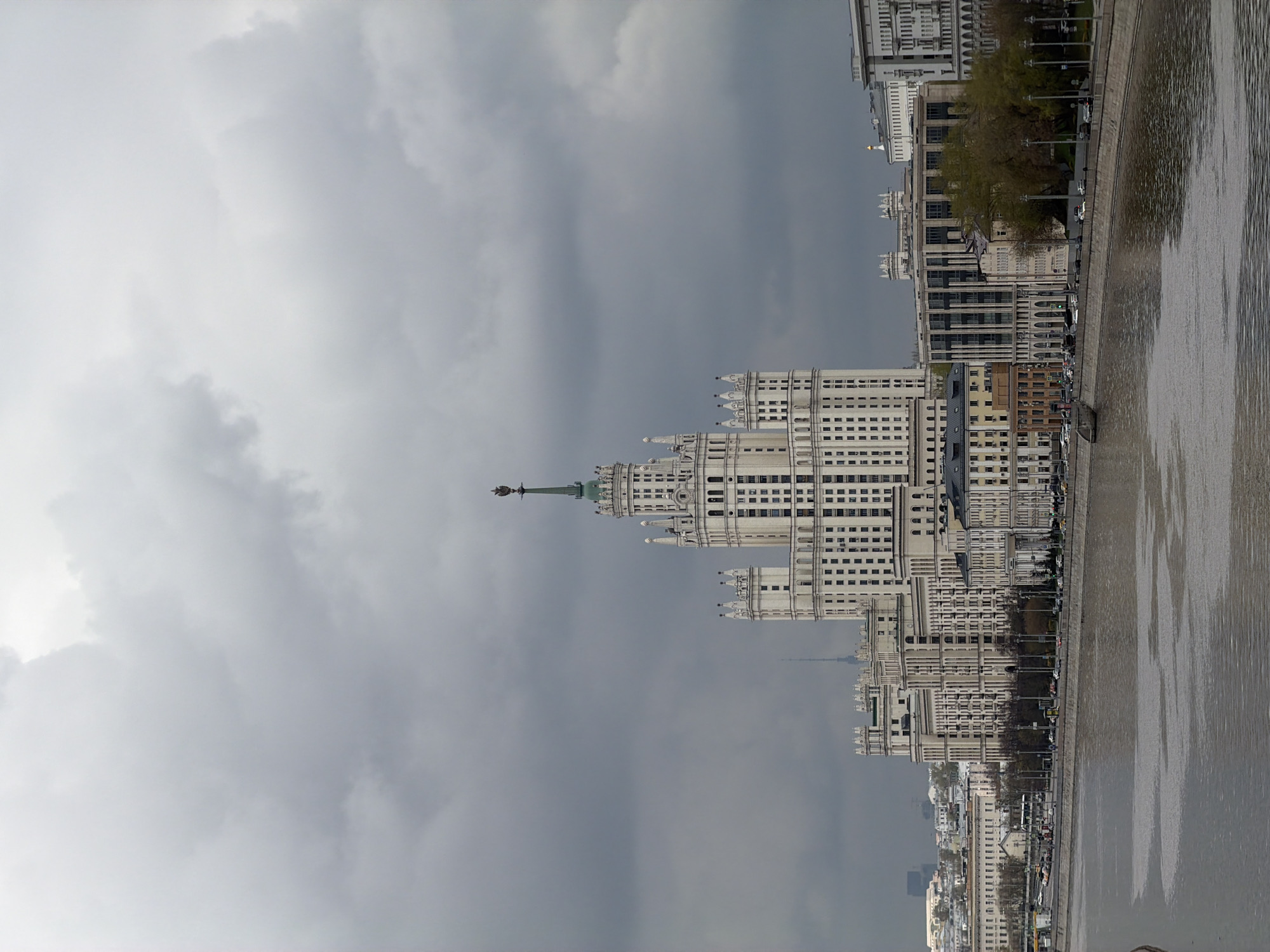 This media file captures a wide view of the Moskva River embankment in Moscow, Russia, under an overcast sky. The image is oriented vertically, showing the river on the right and city buildings on the left.
The dominant subject is a grand Stalinist-era skyscraper, identifiable as the Kotelnicheskaya Embankment Building, with its characteristic tiered structure and a prominent spire topped with a star, dominating the mid-ground on the left. Its light-colored facade contrasts with the dark, heavy clouds above. Many windows are visible across its numerous stories.
Alongside the river, a long stretch of embankment is lined with various multi-story buildings, some featuring classical architectural details like columns and intricate cornices, consistent with Moscow's historical city center. A road or promenade runs along the riverbank, where trees (some with green foliage, others appearing bare) and streetlights are visible. Small, distant vehicles can be glimpsed on the road.
The wide Moskva River occupies the right side of the frame, its surface reflecting the cloudy sky and the distant city structures, creating subtle ripple patterns. The water appears calm, with reflections hinting at the urban landscape.
The overall scene suggests daytime, but the weather is notably gloomy, characterized by thick, grey, low-hanging clouds that cover most of the sky. This creates a muted color palette, primarily in shades of grey, white, and muted greens from the trees. No specific activities or identifiable people are clearly visible, conveying a sense of quiet city life under a somber sky. There is no visible text.