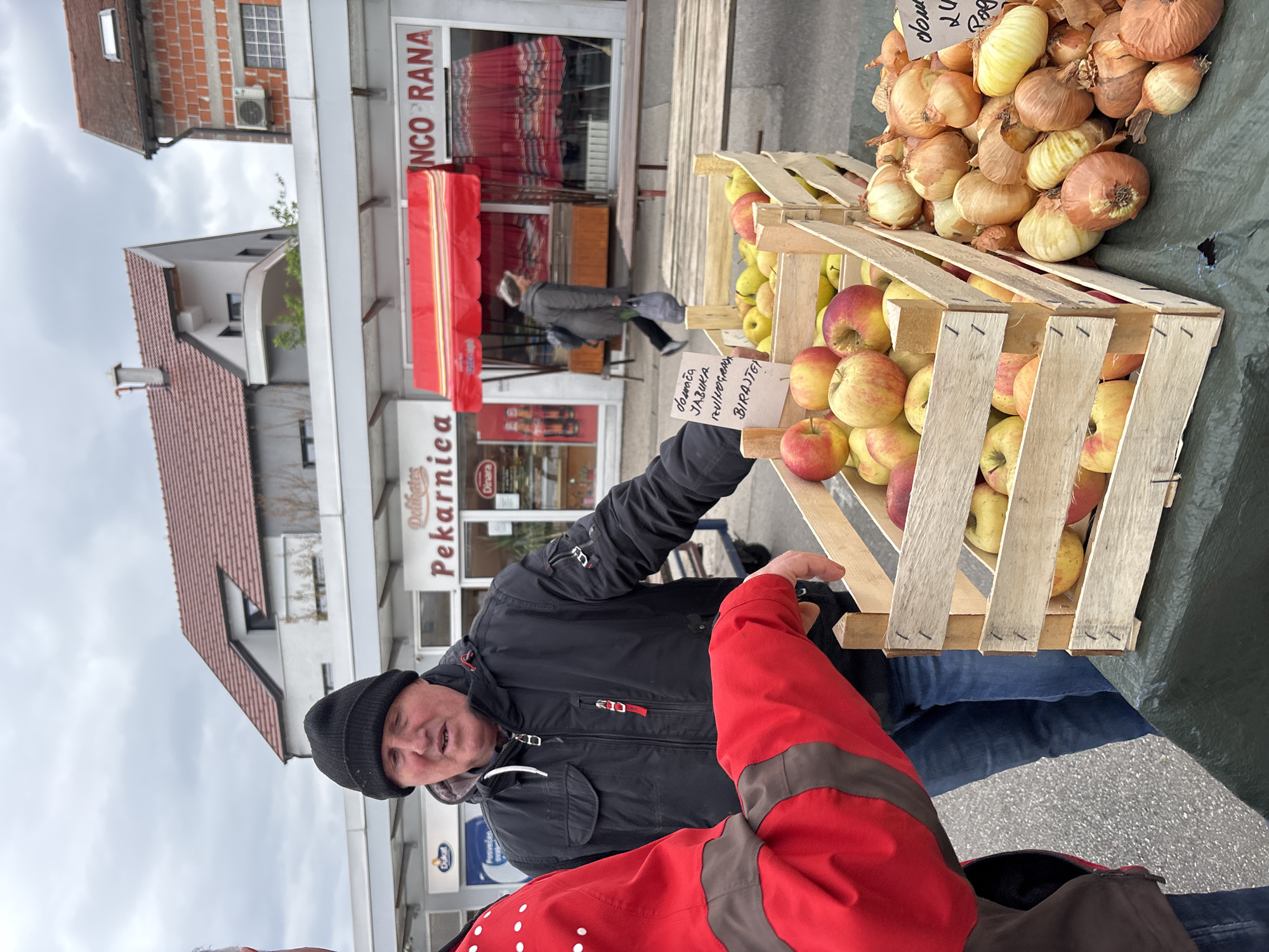 The image captures a street scene in the City of Zagreb, Croatia, showcasing a fruit and vegetable vendor engaged with a customer. The vendor, wearing a black beanie and jacket, stands beside wooden crates filled with apples and onions. He is holding a handwritten sign in Croatian that says "Domaci jabuka poluos, BIRAJTE". A customer, clad in a red jacket with brown shoulder accents, is partially visible. The vendor seems to be gesturing towards the produce, suggesting an interaction related to the sale. In the background, buildings with visible signage, including "Delikates Pekarnica" and "INCO RANA," indicate the commercial nature of the area. Other people are walking in the background. The environment appears to be an outdoor market or street sale.