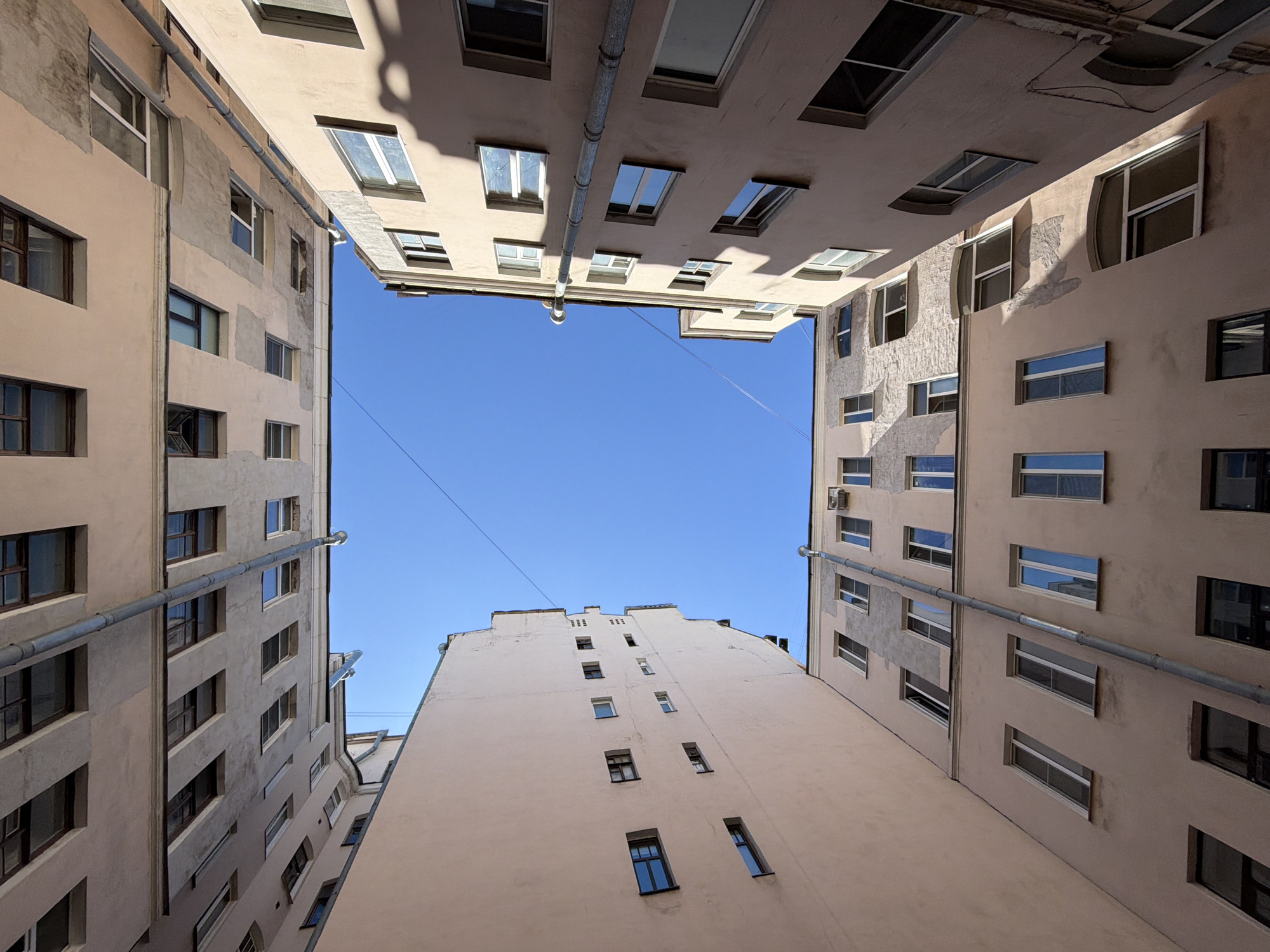 This upward-facing architectural photograph depicts a typical "well courtyard" (двор-колодец) in Saint Petersburg, Russia. Four distinct building facades enclose a rectangular void, revealing a clear, bright blue sky in the center. The buildings are predominantly painted in a faded light pinkish-beige or ochre, with areas of exposed grey plaster, textured surfaces, and peeling paint, indicating wear and repairs, particularly on the upper-right and lower-left structures. Numerous rectangular windows, varying in size and framing styles (some modern white, others older, darker frames), are distributed across the facades. Many windows exhibit reflections of the sky and parts of the opposing buildings. Several grey metal drainpipes are affixed vertically to the exterior walls, notably on the upper-left and lower-right buildings, with one long diagonal pipe visible on the lower-right structure. Bright sunlight illuminates the scene, casting distinct shadows on the building surfaces, particularly on the rightmost building, suggesting a clear, sunny day. A thin black wire or cable traverses the upper-left quadrant of the sky. No people, vehicles, or significant vegetation are present within the frame.
