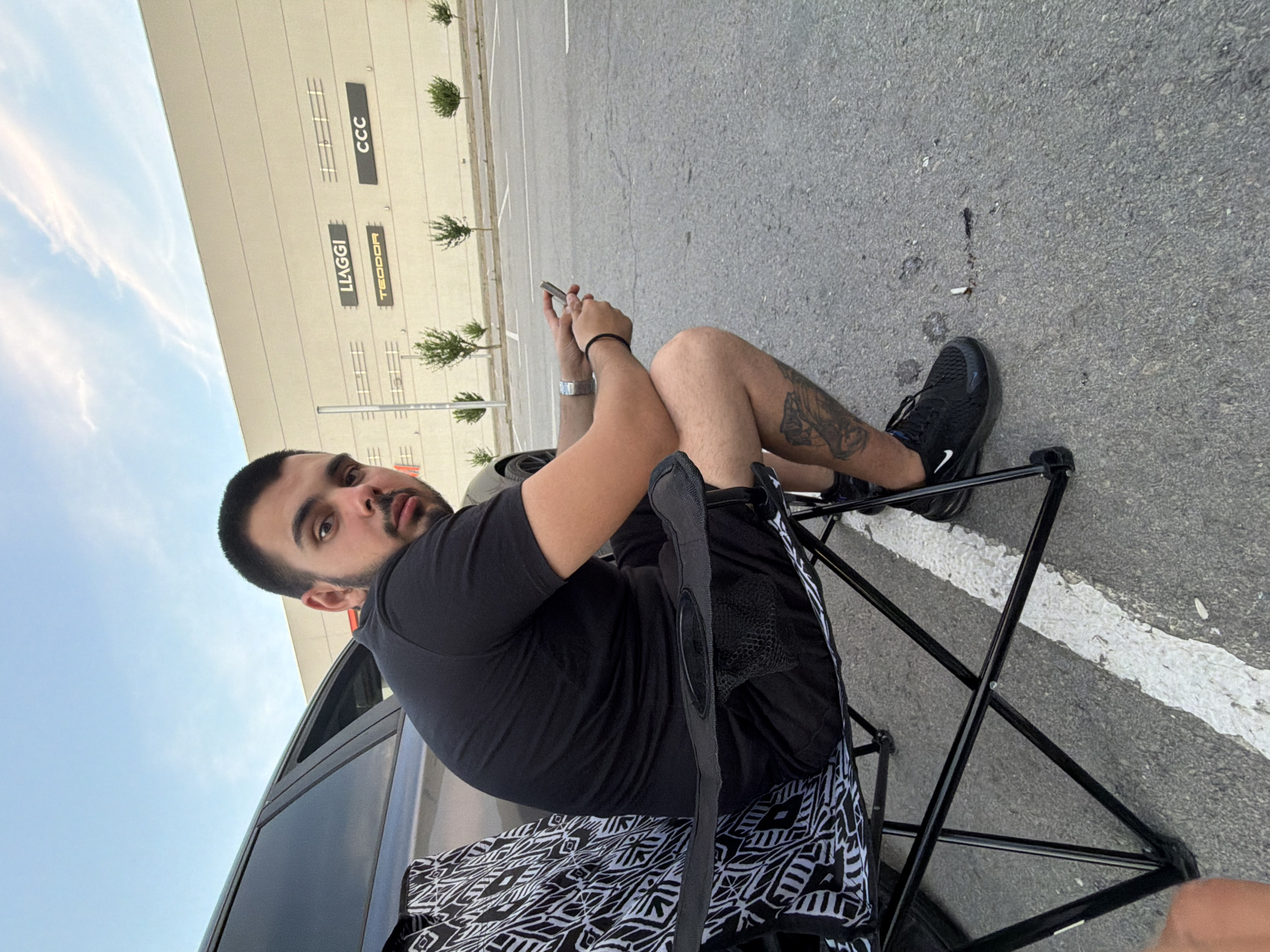 A man with dark hair and a beard is sitting in a black folding chair outdoors, looking towards the camera. He is wearing a black t-shirt and black shorts, with black athletic shoes. The chair is placed on asphalt, next to a vehicle. A patterned blanket with black and white geometric designs is draped over the chair. The man is holding a mobile phone in his hands. He appears to have tattoos on his right leg.
In the background, a light-colored building with signage is visible. The text "LLAGGI" and "CCC" can be clearly read on the building. The ground is a paved surface with white lines, suggesting a parking lot or similar area. Several small, manicured bushes are planted along the building's edge. The sky is a clear blue with some wispy clouds. The scene appears to be set during the daytime, likely late afternoon or early evening given the lighting and the clear sky. The overall impression is a casual outdoor scene, possibly a break or waiting period.