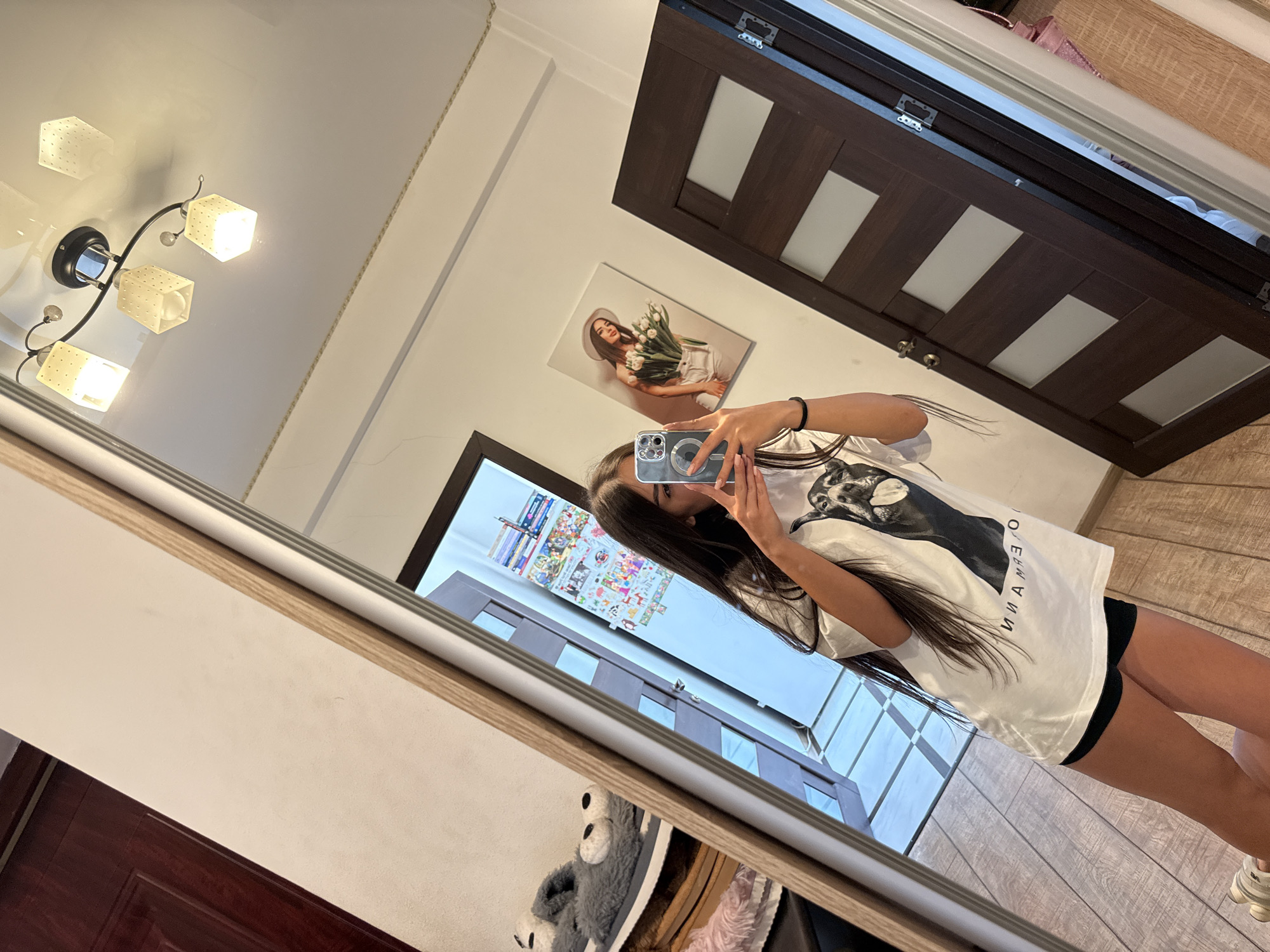 The image captures a young woman taking a mirror selfie in what appears to be a hallway or a room with a large mirror. She is wearing a white t-shirt with a graphic print of a dog and black shorts. Her phone is visible in her hand as she takes the picture. Reflected in the mirror are a wooden door, a ceiling light fixture with multiple lamps, and some wall decorations, including a framed picture of a woman holding tulips. There are also some fuzzy slippers and other items visible to the left in the reflection. The floor is wooden. The lighting is bright, suggesting an indoor setting. There is no explicit information in the image that would allow for the identification of a specific location within Chișinău, Moldova.