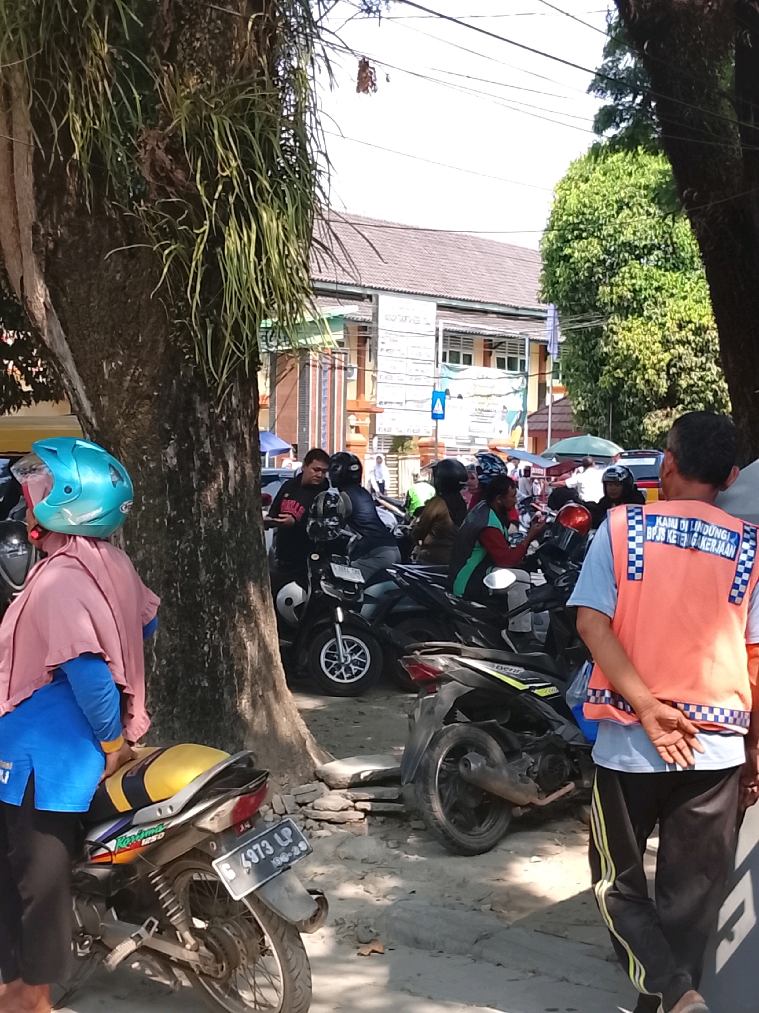 In Slawi, Indonesia, a group of people are gathered near a street lined with trees. A woman in a pink headscarf and blue shirt stands next to a motorcycle with a license plate that reads "G 4973 LP." Behind her, a man wearing an orange vest with checkered blue and white trim faces away from the camera, his hands clasped behind his back. Several other individuals are seen on motorcycles in the background, some wearing helmets. A building with a brown roof is visible behind the people, suggesting an urban or semi-urban setting. The ground is a mixture of dirt and pavement, with exposed tree roots and some scattered stones. The overall scene appears to be a casual street gathering or a moment of pause in a busy area.