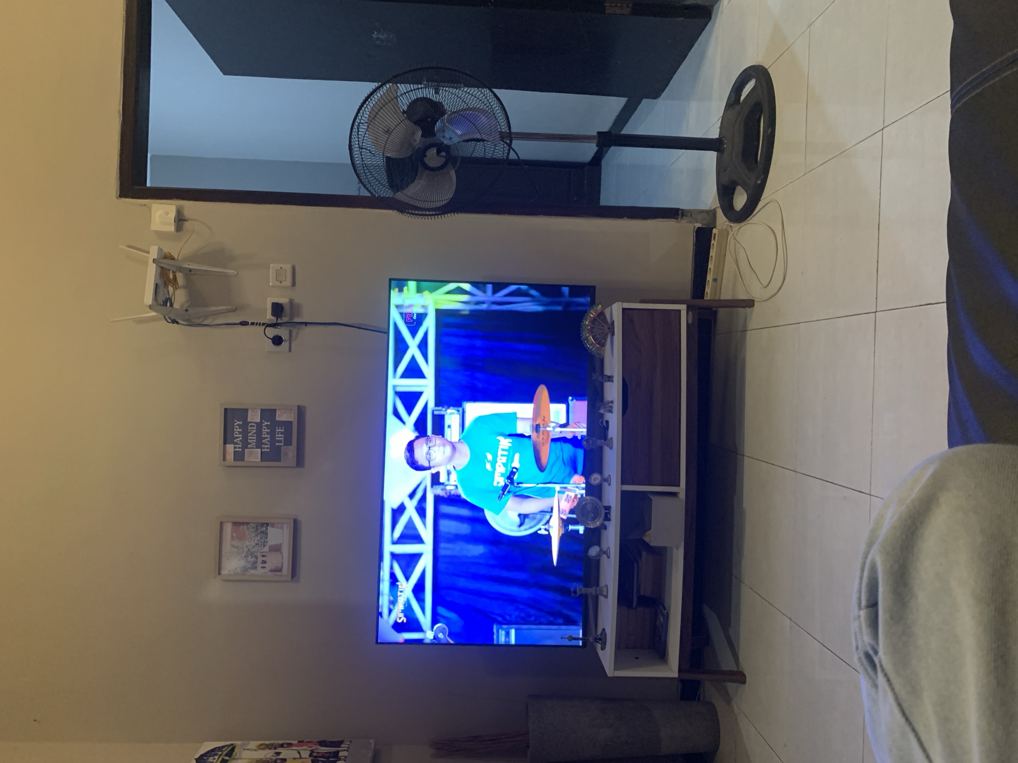 The image depicts an indoor scene, likely a living room in Malang, Indonesia. A large flat-screen television is the central focus, displaying a man in a teal t-shirt playing drums on a stage with a blue backdrop. To the left of the television, there is a tall, cylindrical gray object. Above the television, mounted on the wall, are two framed pictures. The left frame contains a floral motif, while the right frame has text reading "HAPPY MIND HAPPY LIFE". Further up and to the right, a white Wi-Fi router with two antennas is attached to the wall. A black standing fan is positioned to the right of the television, partially obscuring a dark doorway. The floor is tiled with white squares. A portion of a gray blanket or clothing item is visible in the foreground, suggesting the photo was taken from a seated position. Electrical outlets and cables are visible on the wall near the fan and router. A white media console with wooden drawers sits beneath the television, holding various small decorative items and storage boxes.