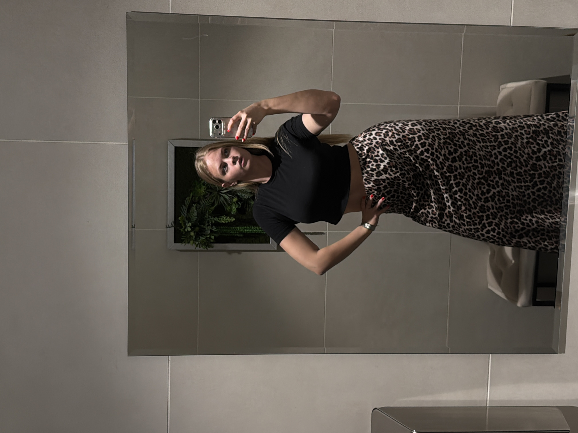A fair-skinned woman with blonde hair is taking a mirror selfie. She is wearing a black crop top and a long leopard-print skirt. The environment appears to be a bathroom with light-colored tile walls. There is a framed artificial plant on the wall behind her. A toilet and hand dryer are visible in the background. The location in Izhevsk, Russia cannot be determined from the image.