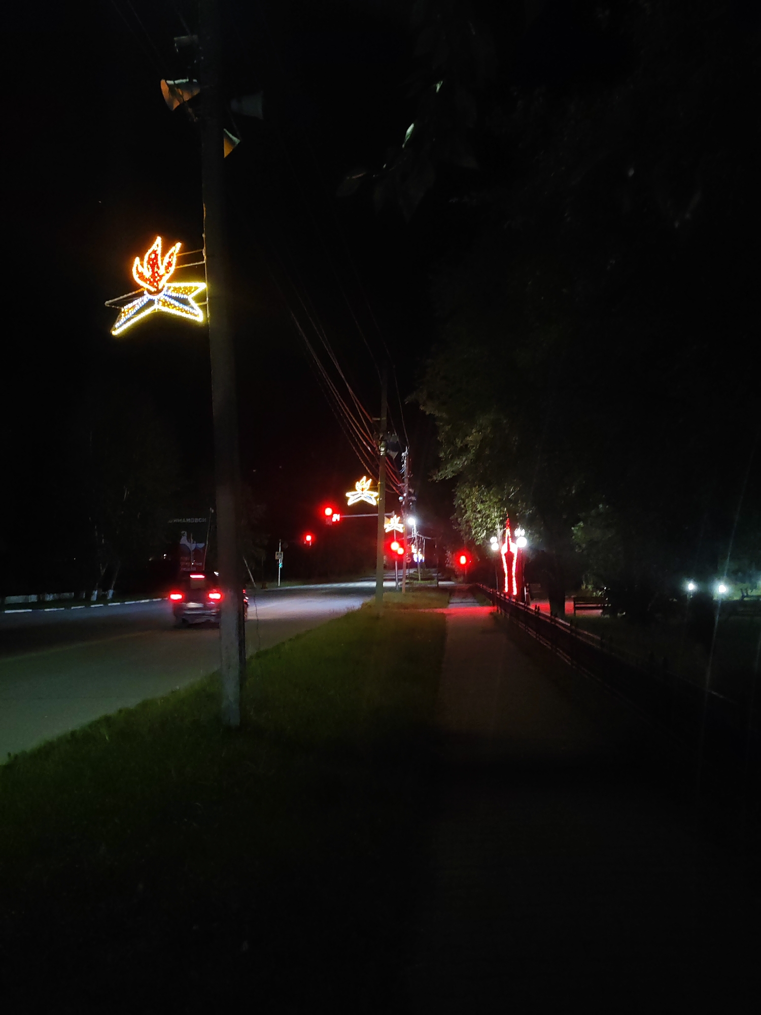 The image depicts a nighttime scene along a road in Shimanovsk, Russia, with decorative lights illuminated. On the left side of the road, a dark-colored car is driving away from the camera. The road itself is illuminated by the car's headlights and what appear to be streetlights. Along the right side of the road, a sidewalk or pedestrian path is visible, bordered by a dark railing and trees. Decorative lights, shaped like stars or stylized flames, are strung on poles along the road, casting a warm glow. Traffic signals displaying red lights are also visible. The overall environment appears to be an urban or suburban area, possibly a park or a street lined with trees, during the evening or night.