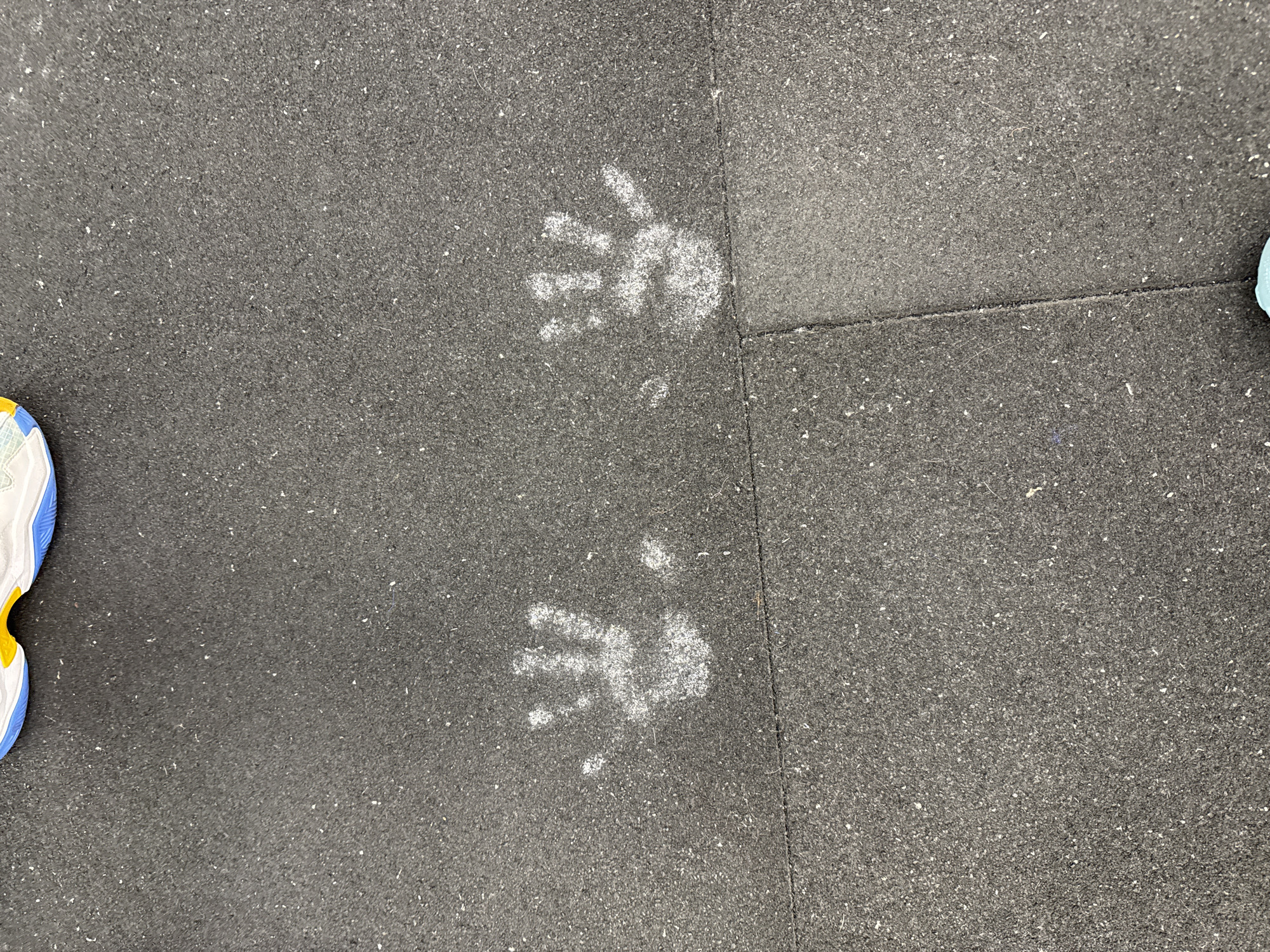 The image shows two handprints in white powder on a dark gray, textured floor. The prints appear to be from a child's hands. A section of a shoe with white, yellow, and blue accents is visible on the left edge of the image. Another shoe can be seen at the far right. The flooring is divided by a seam. The location cannot be determined from the image.
