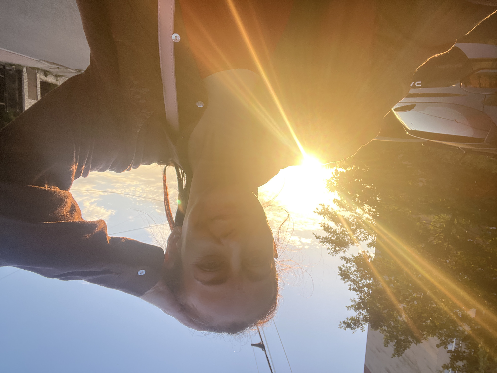 The media file is a slightly overexposed, vertically oriented selfie of a person in an outdoor setting. The person is facing the camera, with their head tilted downwards. The bright sunlight is directly in front of them, creating lens flare and washing out details in the center of the image. The person appears to be wearing a dark, collared shirt with light-colored trim and a button. Their hair is dark and has some wispy strands catching the light.
The background is blurred due to the bright light and shallow depth of field. To the left, there's a glimpse of a building with a light-colored facade and some windows. To the right, there are trees with green foliage and more sunlight streaming through the branches. In the middle distance, the rear of a light-colored car is visible, with the word "CIVIC" written on the trunk. The sky is clear and blue.
The strong sunlight and the angle of the sun suggest it is late afternoon or early morning, likely during a golden hour period. The overall mood is warm and bright, though the overexposure makes it difficult to discern specific emotions. The setting appears to be urban or suburban, possibly a street or driveway. No other people are visible.