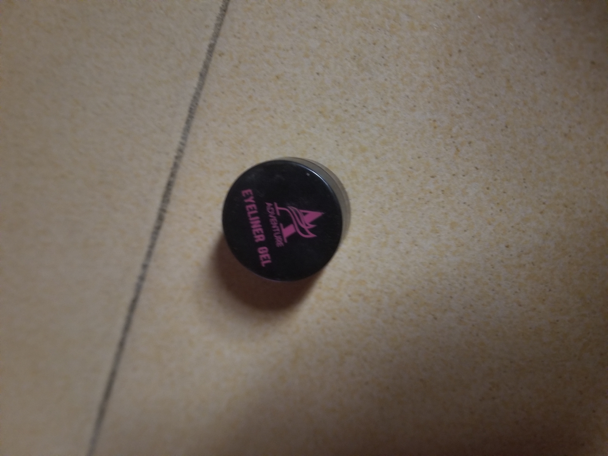 A black pot of eyeliner gel with pink text and a pink logo sits on a textured, light-colored surface. The text on the pot reads "EYELINER GEL" and "ADVENTURE." The background surface appears to be a floor or countertop with a subtle speckled pattern. The image is lit from above and to the left, casting a shadow to the right of the pot. There is a thin, dark line running diagonally across the upper portion of the image, possibly a crack or seam in the surface. The scene is indoors, with no indication of its specific location within Jauro Yinu, Nigeria. There are no people or animals present in the image.