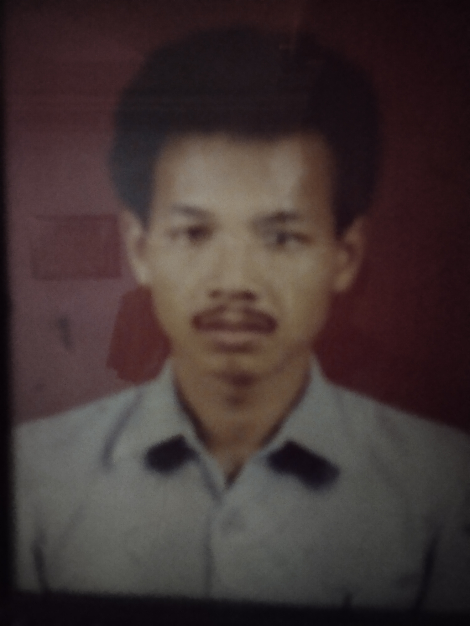 A faded, grainy portrait shows a man from the chest up against a dark red, textured background. The man has short, dark hair, a thin mustache, and is wearing a light-colored collared shirt. There are two dark rectangular shapes visible on the shirt, possibly epaulets or patches. The lighting is dim and casts soft shadows, contributing to the overall aged appearance of the photograph. The specific location in Dermasandi, Indonesia, is not discernible from the image itself.