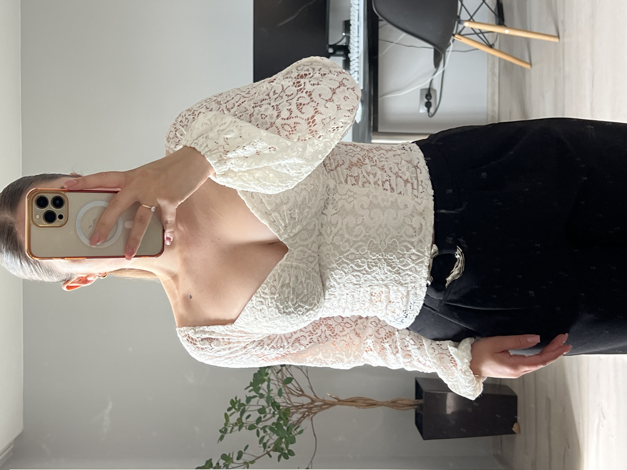 A woman is captured taking a mirror selfie indoors, with the image rotated 90 degrees clockwise. She holds a gold/rose gold iPhone, featuring multiple camera lenses and a visible MagSafe ring, in her left hand. A diamond ring is worn on her left ring finger.
She is dressed in a white, long-sleeved lace top with a deep V-neckline and ruffled cuffs, paired with black high-waisted pants or a skirt. The intricate floral lace pattern of her top is clearly visible. Her other hand rests casually near her hip.
The setting is a modern indoor space, likely in Ternopil, Ukraine. The background reveals a contemporary chair with wooden legs and a black seat, part of a desk or table with computer equipment (monitor and keyboard are partially visible), and a power outlet on a light-colored wall. Below the mirror, a decorative potted plant with thin, twisted branches and green leaves is placed in a dark, rectangular planter. The floor, which appears to be reflective, shows a partial reflection of the plant. The lighting suggests it is daytime indoors. No discernible text is visible in the image.