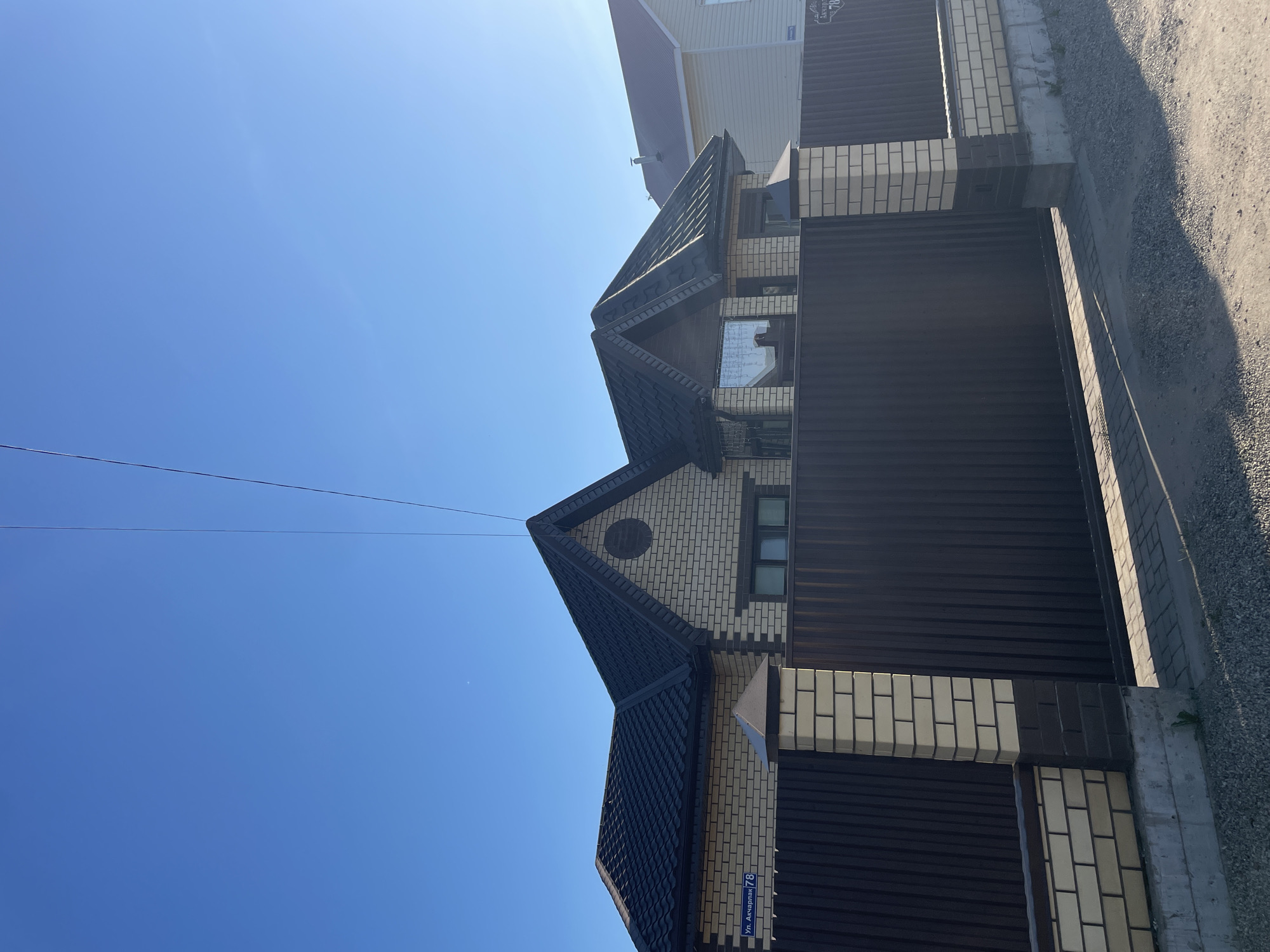 The image shows a building exterior against a clear blue sky. The building features a dark-tiled roof, tan brick walls, and a dark brown metal fence. On the building is a street sign reading "ул. Акчарлак, 78", indicating the location as Ulitsa Akcharlak, 78 in Kazan, Russia. There are no people visible in the image. The environment suggests a residential area.