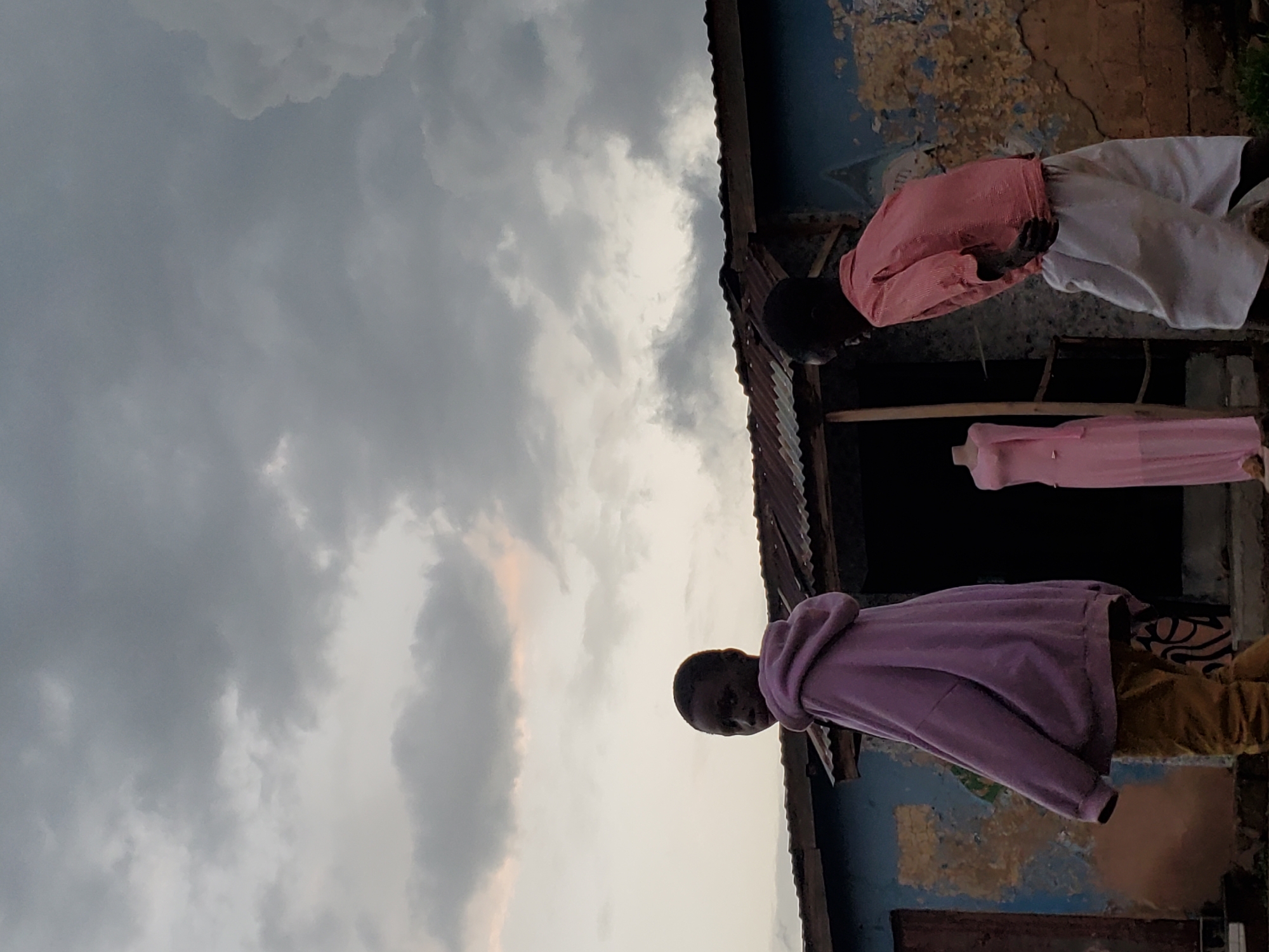 The image, taken in Jos, Nigeria, shows an outdoor scene with a cloudy sky. In the foreground, a child wearing a purple hoodie and yellow pants is looking towards the camera with a neutral expression. Behind this child, another person, possibly an older child or adult, is wearing a pink shirt and white pants. A pink dress is displayed on a mannequin or hanger in the background, suggesting a potential clothing store or tailor's shop. The setting appears to be an urban or semi-urban area, with corrugated iron roofing and a weathered building wall visible. The time of day is likely late afternoon or early evening, given the overcast sky and soft lighting. The overall mood is subdued, possibly due to the weather. There is no visible text in the image.