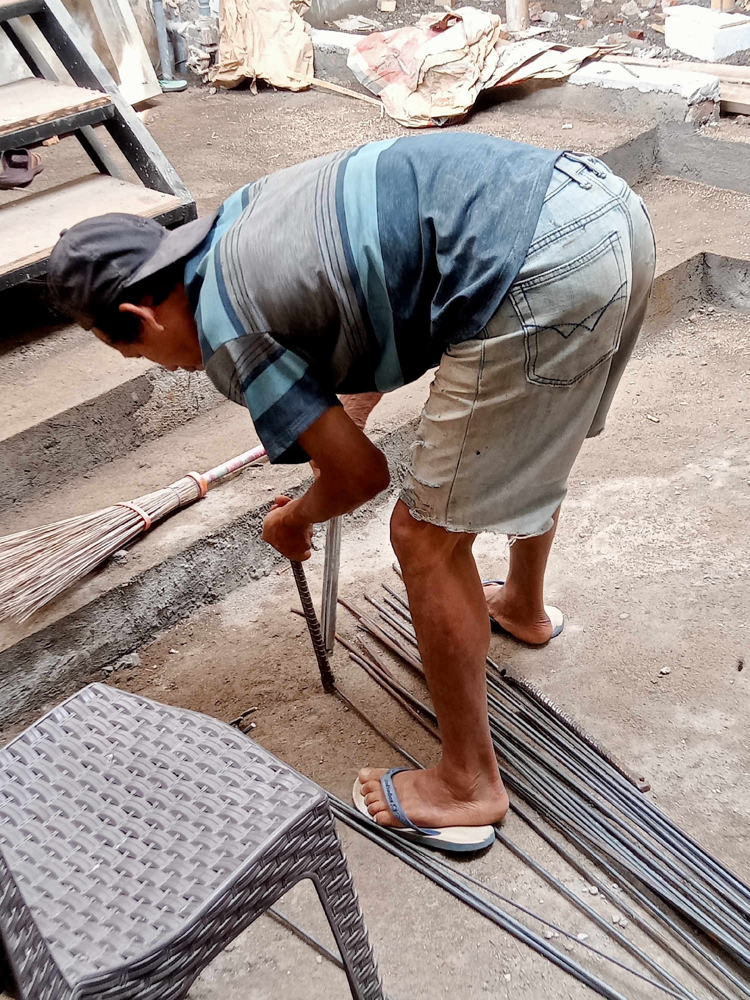 An Indonesian construction worker is bending over, holding a metal rod in his right hand, at a construction site in Simo, Indonesia. He is wearing a blue and grey striped t-shirt, light blue denim shorts that are torn at the hem, a black baseball cap, and blue and white flip-flops. A broom made of natural fibers is lying on the ground to his left, next to a concrete step. Several metal rods are scattered on the ground to his right. In the foreground, a grey woven plastic stool is partially visible. The background shows construction materials, including bags of cement and what appears to be building debris.