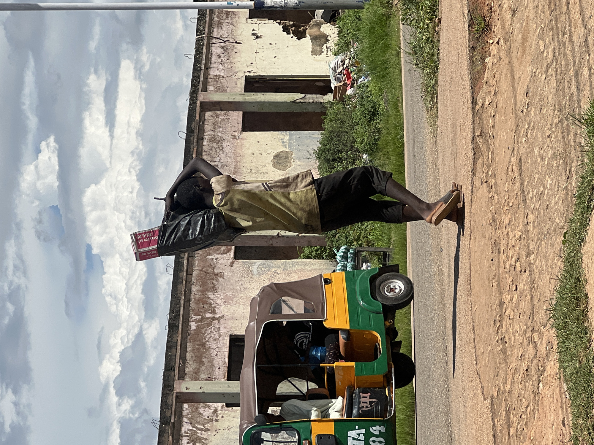 A young boy carries a large black trash bag over his shoulders, with a pink and red box peeking out from the top. He wears a yellow t-shirt and dark shorts. The boy walks on a dusty path beside a road. To his left, a green and yellow auto-rickshaw is parked. In the background, a dilapidated building with crumbling walls and doorways stands under a cloudy sky. Greenery and debris are visible around the building. The scene appears to be in Jos, Nigeria.