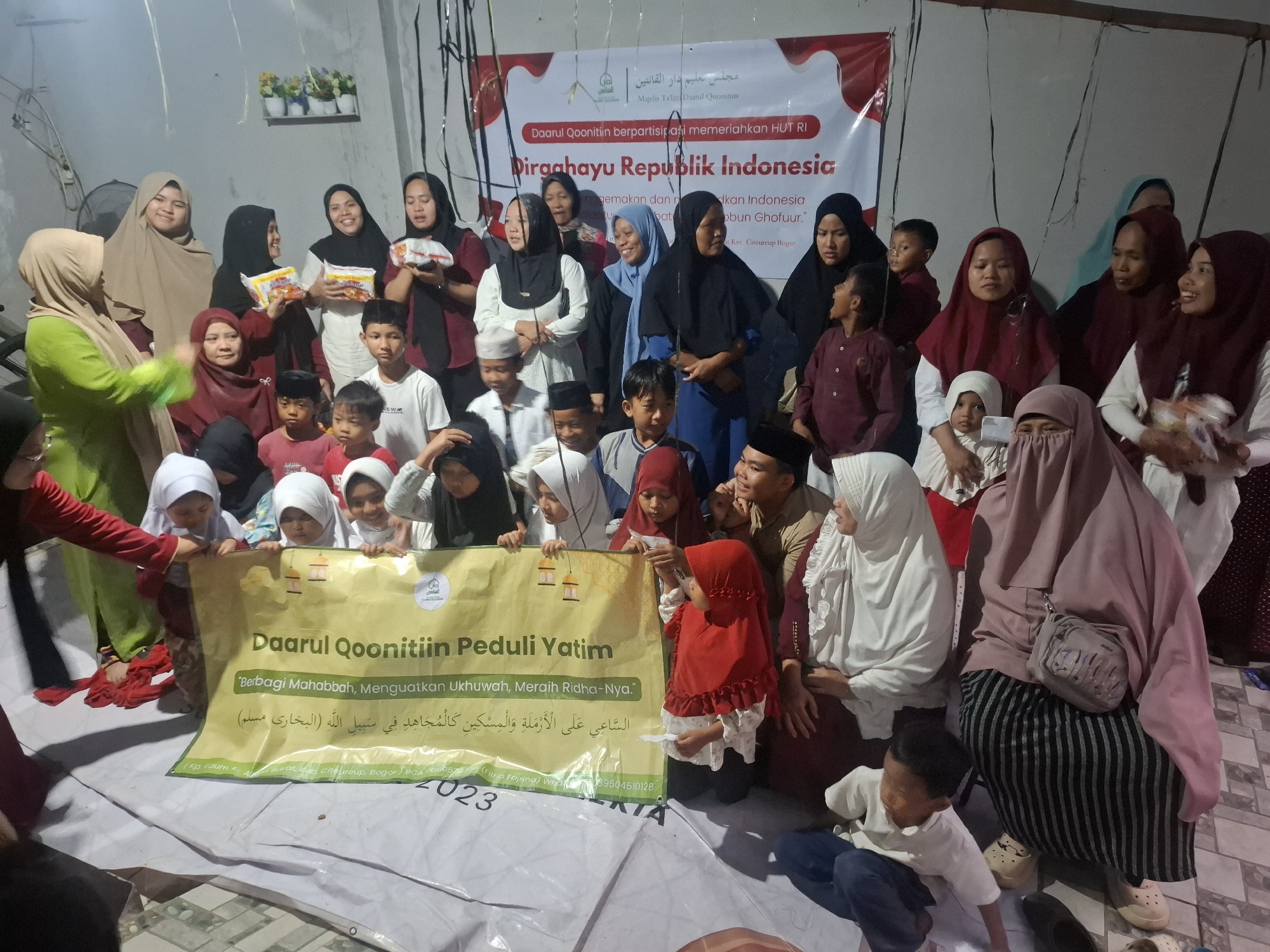 The image depicts a gathering of people, predominantly women and children, indoors. A banner in the background, partially obscured by hanging streamers, reads "Daarul Qoonitiin berpartisipasi memeriahkan HUT RI" and "Dirgahayu Republik Indonesia". The text on the banner also includes Arabic script and mentions "Majelis Ta'lim Daarul Qooniim" and "Kec. Citeureup, Bogor". This suggests the event is taking place in or around Citeureup, Bogor, Indonesia, which is located in the city of Babakan Madang.
The group is gathered around a yellow banner that says "Daarul Qoonitiin Peduli Yatim" and "Berbagi Mahabbah, Menguatkan Ukhuwah, Meraih Ridha-Nya." Below this, there is Arabic text and the year "2023".
Many individuals are wearing hijabs and traditional clothing. Some are holding small white papers or items. A few young children are prominent in the foreground, some seated and others standing, looking towards the camera or at the banner. The environment appears to be a simple indoor space, possibly a community hall or a large room, with bare walls and a visible wooden beam structure at the top. There are also some decorations, including streamers. The lighting is somewhat dim, creating a muted atmosphere.