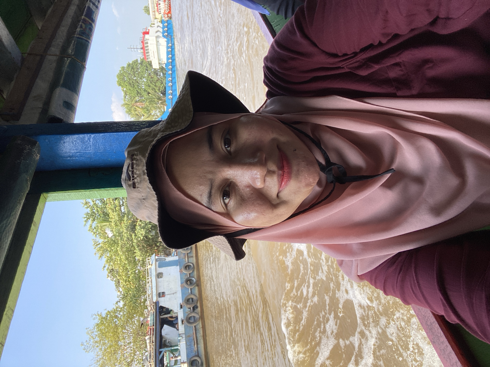 A woman wearing a pink hijab and a maroon shirt, along with a brown bucket hat, is taking a selfie on a boat. She is smiling and looking at the camera. The boat is moving on a body of water, creating ripples and waves. In the background, several other boats are visible, as well as lush green trees and a clear blue sky. The scene suggests a journey or transport on the water, possibly in a tropical or subtropical region. The location is Berangas Timur, Indonesia.