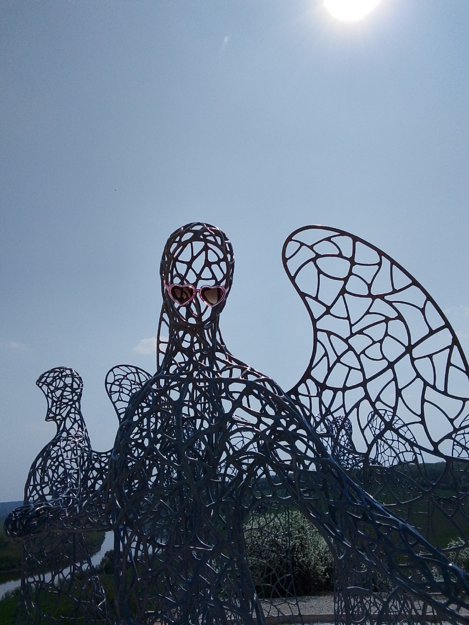 The image depicts three metal sculptures of human figures with wings, standing outdoors under a bright, sunny sky. The figures are constructed from interconnected metal pieces, creating a web-like appearance. The central figure is wearing pink heart-shaped sunglasses. In the background, there's a view of a river and green vegetation, suggesting a location overlooking a natural landscape. The scene is in the city of Каменка, Russia.
