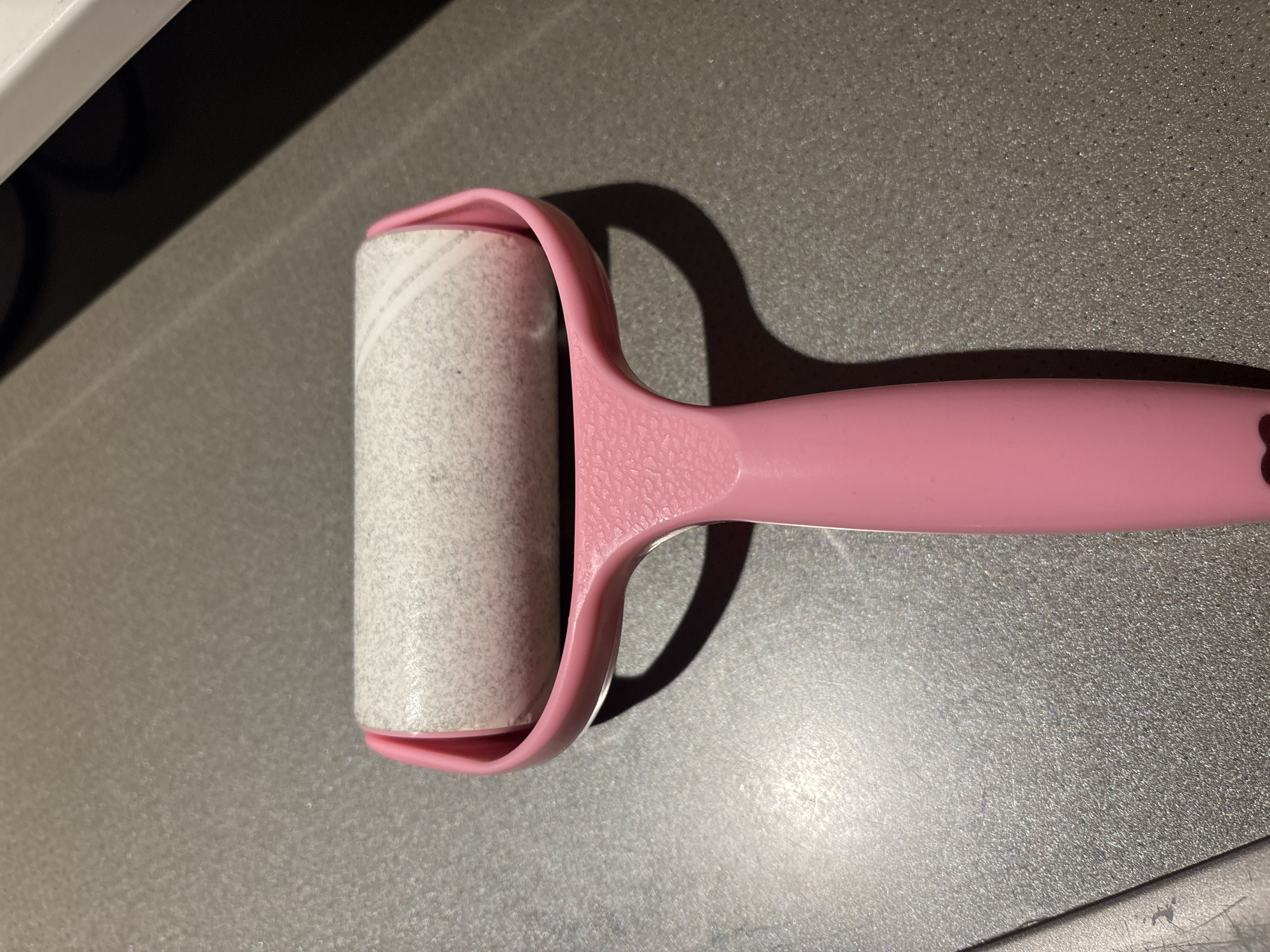 A lint roller with a pink handle and a white roller is positioned on a speckled gray countertop. The roller appears to be made of a sticky material and has a textured surface. The handle has a heart-shaped cutout near the bottom. The scene is indoors, and there are no people present. The lighting suggests an indoor setting, possibly a home or office, in Dubai, United Arab Emirates.