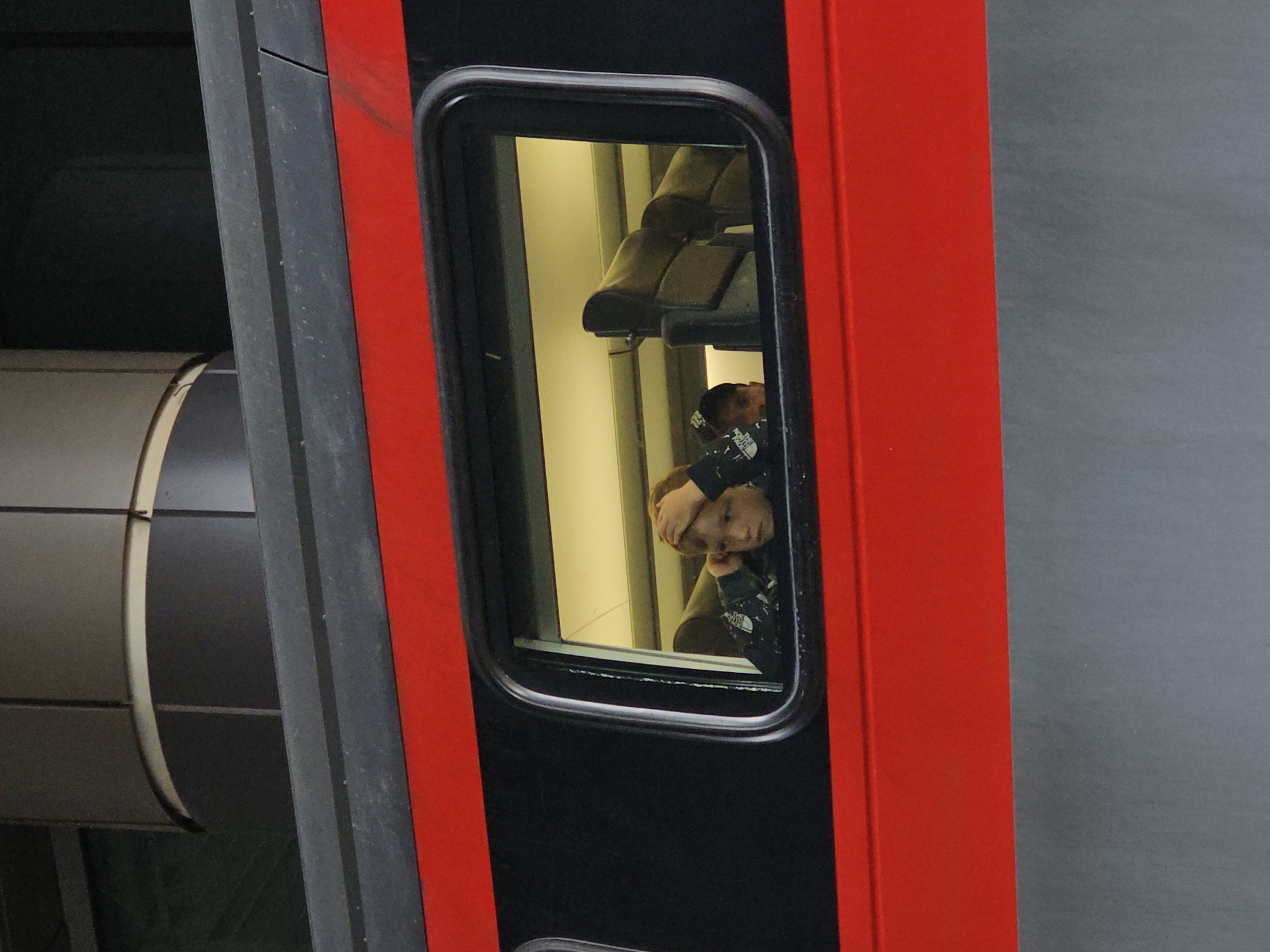 The image captures a close-up view of a train window. Inside the train, a young boy with blond hair is visible, resting his head on his hand and looking out of the window. He is wearing a dark-colored jacket with a white graphic on it. Another child, possibly younger, is partially visible behind him, wearing a baseball cap. The interior of the train carriage is illuminated by overhead lighting, revealing the backs of seats. The train's exterior features a distinctive red stripe running along its side, above the window. The train is stationary, and the background outside the window is blurred, suggesting it is either at a station or moving slowly. The image does not provide any specific visual cues that would definitively place it in the city of Trekhgorka, Russia, or any other urban location.
