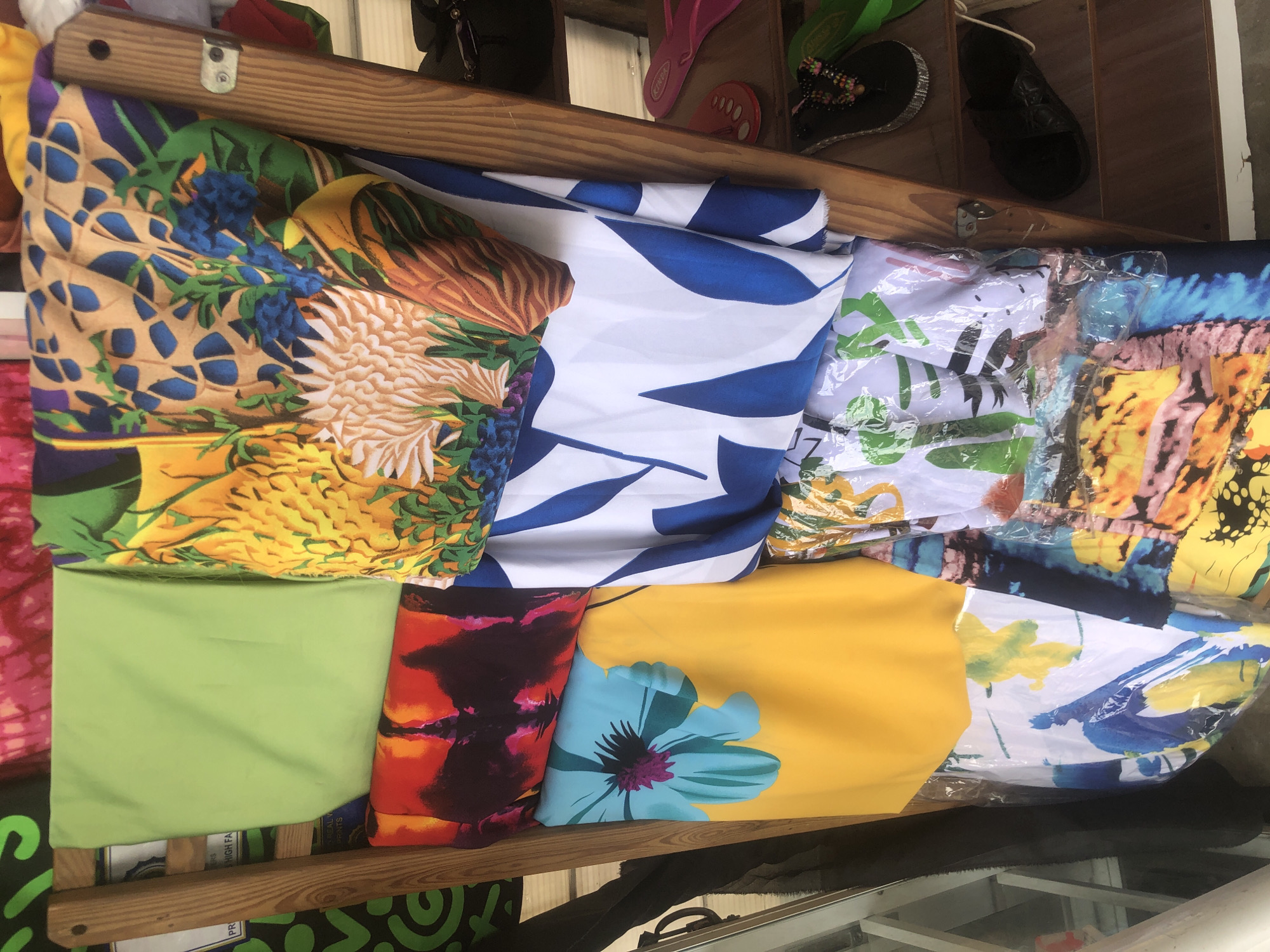 The image presents a vibrant display of folded textile products, likely swimwear or beach attire, neatly arranged on a wooden shelving unit in what appears to be a market stall in Ado, Nigeria.
The top row of the display features several distinct patterns. On the left, a fabric exhibits a detailed design of pineapples, blue berries, and green tropical leaves against a mottled brown and beige background. Adjacent to it is a piece with striking, large blue abstract shapes set against a crisp white background. Further right, a textile with green palm leaf patterns is visible through clear plastic packaging. The item on the far right of this row shows an abstract, colorful print in shades of yellow, blue, and pink.
The bottom row continues the bright assortment. To the left is a solid, plain lime green fabric. Next to it, a piece features a dynamic blend of dark and vibrant orange-red patterns. Following this is a bright yellow fabric adorned with a large, light blue tropical flower with a purple center. The rightmost item in the bottom row, also encased in clear plastic, displays a white background with yellow and blue abstract motifs.
On the top wooden shelf, above the textile display, various pieces of footwear are visible. These include a pair of black slide sandals, a black flip-flop embellished with colorful beads, and a pink flip-flop where the brand name "KERIEA" can be faintly discerned. The overall scene suggests a casual retail environment, characteristic of a local market setting. Partially visible text on a label below the lime green fabric shows fragmented words like "S HIGH FA" and "PR".