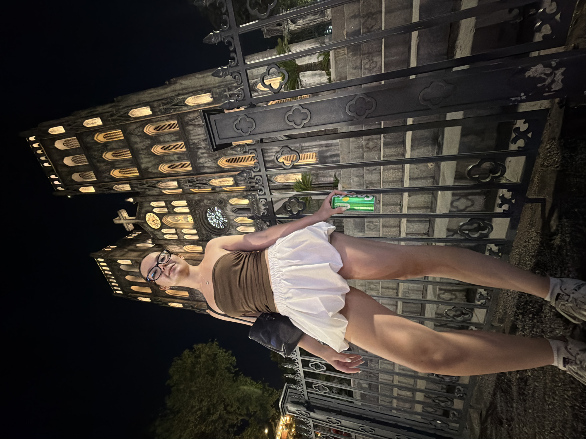 A young woman stands in front of a building with Gothic architecture at night. She is wearing a brown strapless top, a white skirt, glasses, and sneakers. She is holding a can of what appears to be a beverage in her right hand. The building is illuminated, highlighting its ornate windows and stonework. A metal fence with decorative elements is in the foreground. Trees are visible in the background, and the sky is dark, indicating it is nighttime. The scene suggests a casual outing in a historically significant or visually striking location, possibly in Hanoi, Vietnam given the context.
