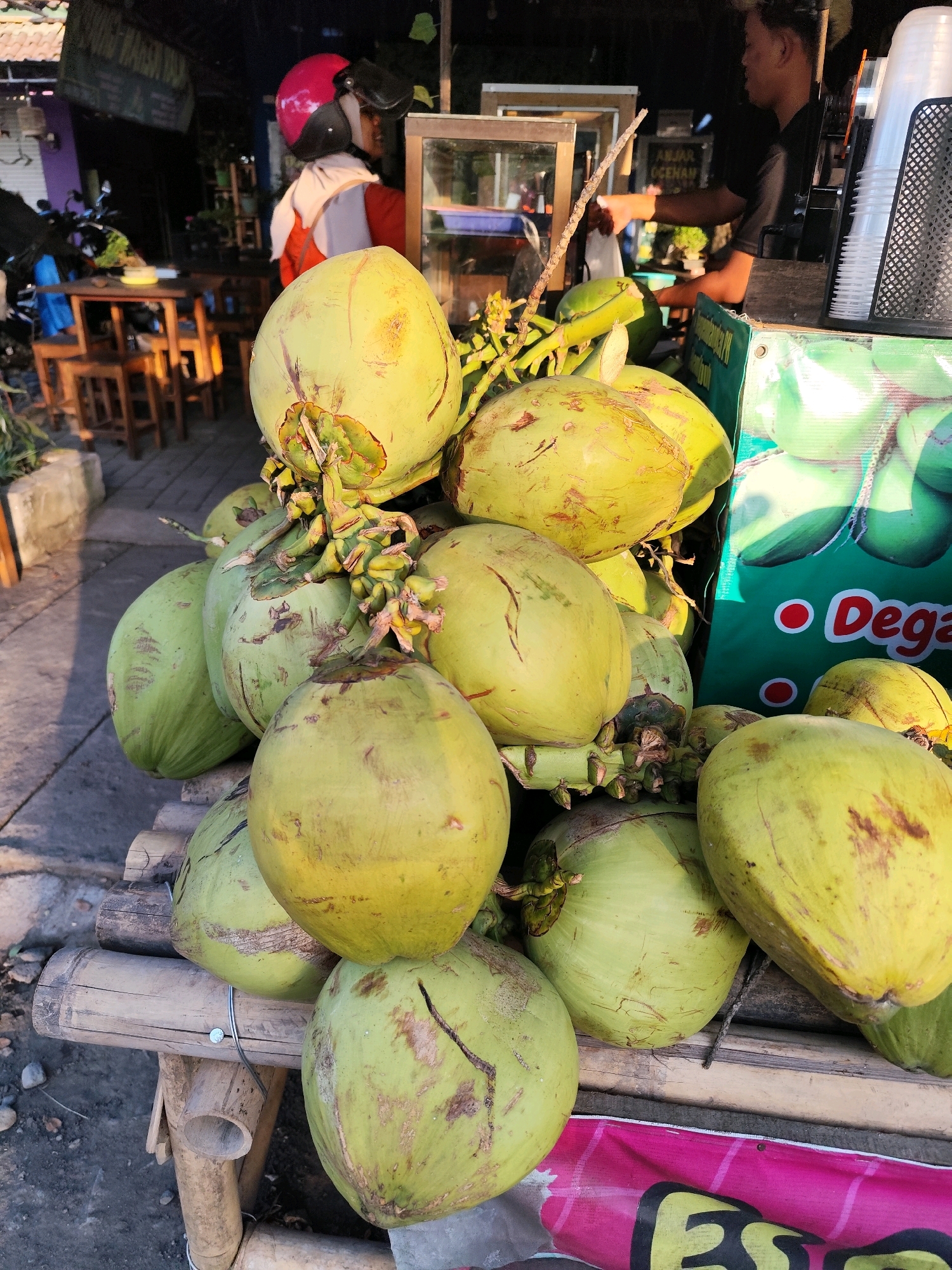 The image shows a pile of young coconuts displayed on a bamboo stand in an outdoor market setting. In the background, a person wearing a pink helmet and a white scarf is visible, as well as a vendor interacting with a customer at a stall. The stall appears to be selling drinks, as indicated by stacked plastic cups and a green banner with red circles and white text that reads "Deg". The ground is paved with concrete and small stones. The overall atmosphere suggests a lively market scene in a tropical location. The image appears to be taken in Bambanglipuro, Indonesia, given the textual cues of "Deg" possibly relating to "Degan" (Indonesian for coconut) and the general appearance of the environment.