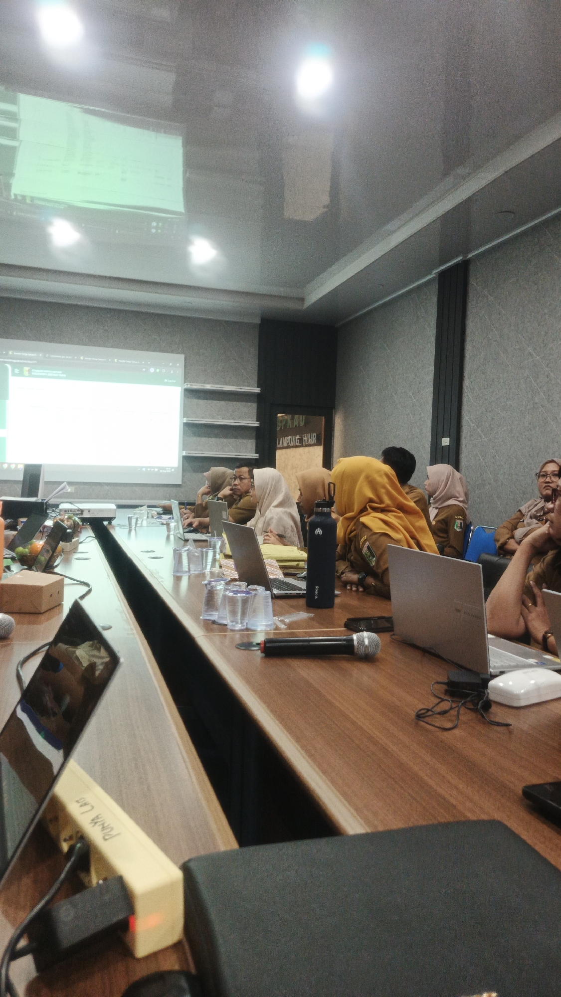 A meeting is taking place in a modern conference room, likely in Sukadana, Indonesia, as indicated by a sign reading "SPKAU LAMPUNG TIMUR" visible on the wall. The room features a large, polished wooden conference table with multiple laptops, microphones, and glasses of water spread across it. A projector is set up, projecting a presentation onto a screen at the far end of the room. Several individuals, predominantly women wearing hijabs and uniforms or work attire, are seated around the table, engaged with their laptops or listening to the presentation. One person in a bright yellow hijab is prominent in the center of the image. The ceiling is reflective, showing the glare of overhead lights. The overall atmosphere suggests a professional gathering or training session.
