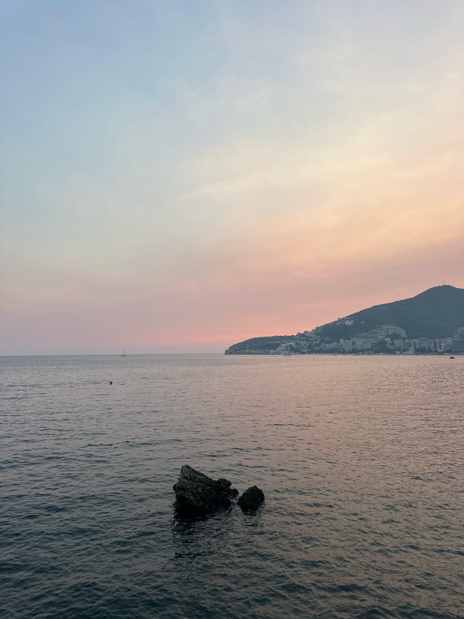 The image depicts a serene seascape with a pastel-colored sky at dusk. The sky transitions from a pale blue at the top to soft pink and orange hues near the horizon. The calm sea reflects these colors. In the foreground, a cluster of dark rocks emerges from the water. Further out, a small sailboat is visible. Along the distant coastline, the city of Budva, Montenegro, is nestled against a dark, forested hill. Buildings of various sizes and colors are scattered across the hillside, and some structures are visible along the waterfront. The overall impression is one of tranquility and the end of a day.