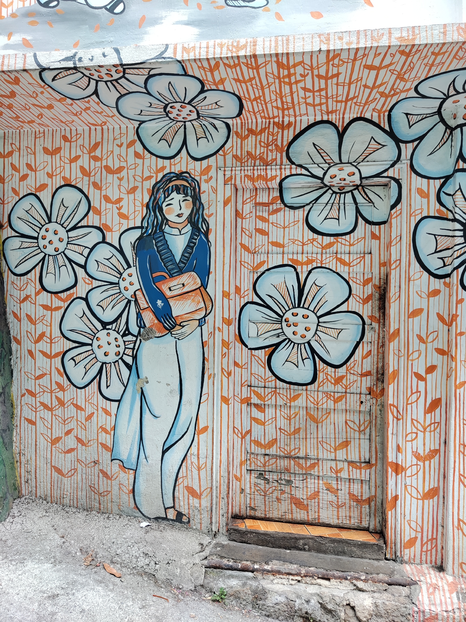 A mural in Da Lat, Vietnam, depicts a woman standing to the left of a painted door. The woman has long blue hair and wears a blue jacket over a white dress. She carries a brown bag. The background consists of orange leaf patterns on vertical white lines and large blue flowers. The mural is on a wall with a visible corner, suggesting an alleyway or building entrance. A concrete ground is visible at the bottom of the image, leading up to the painted door.